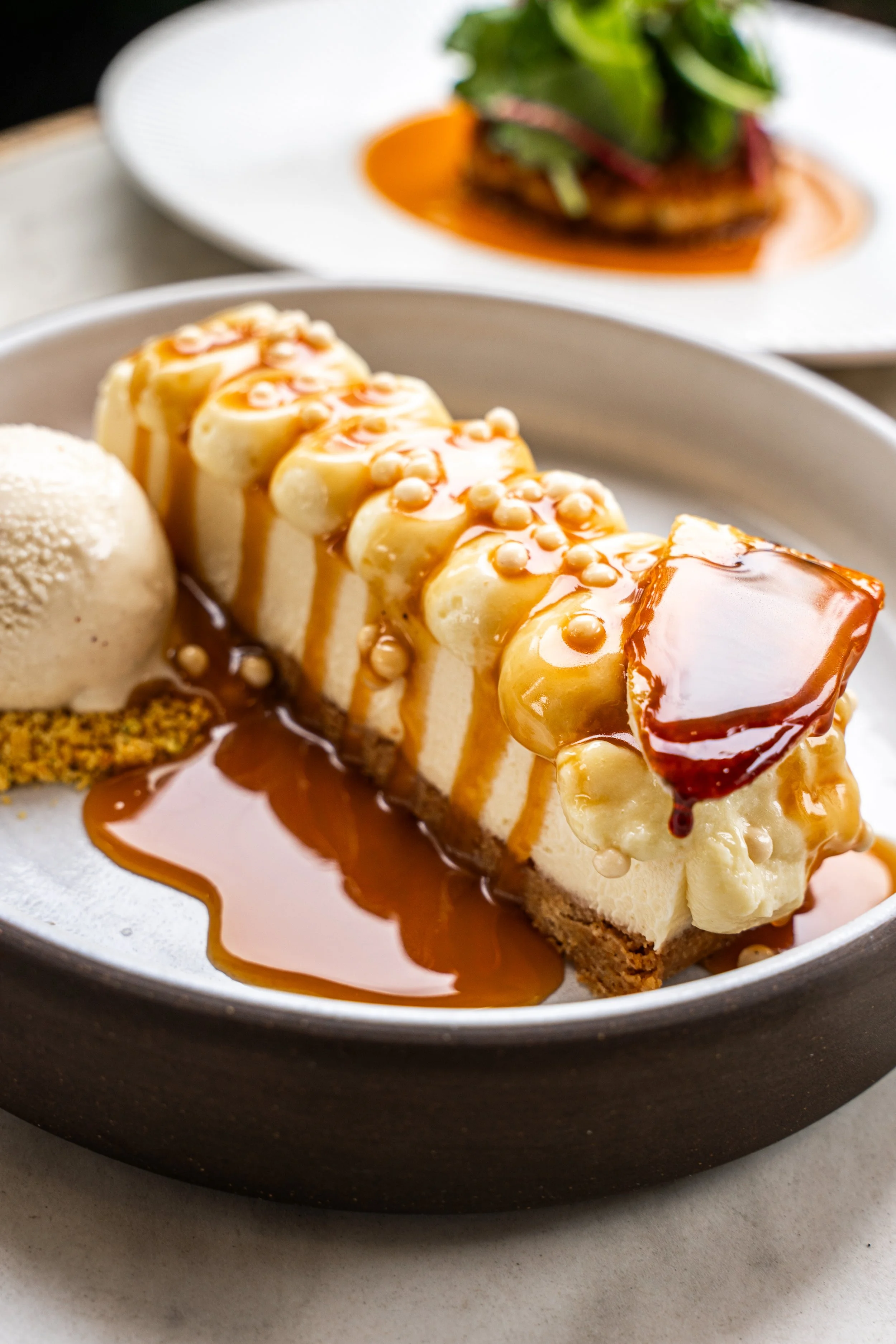 Cheesecake topped with caramel sauce, pearls, and a caramelized slice, served with a scoop of ice cream on the side.