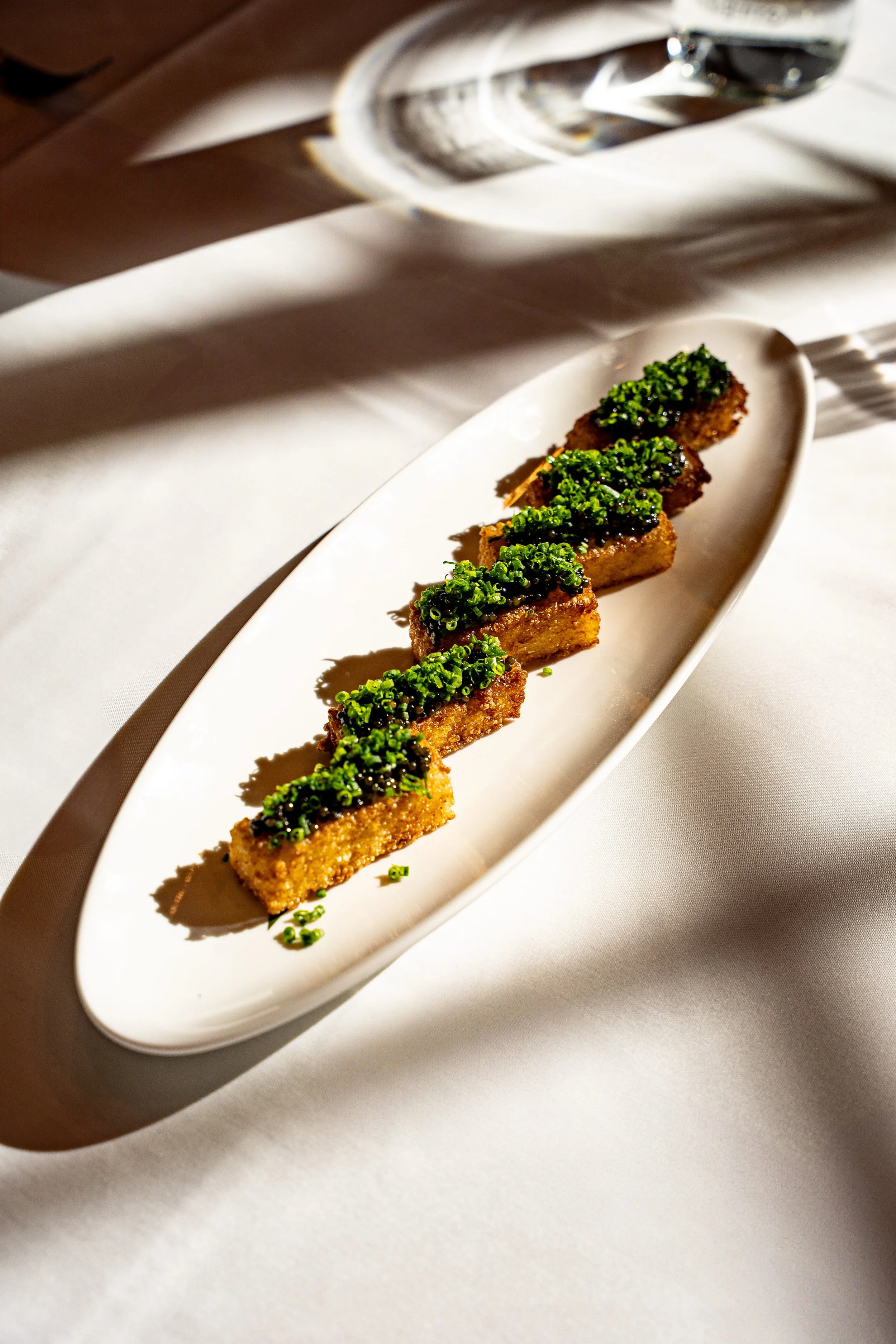 Five pieces of fried food topped with chives on a white oval plate, on a white tablecloth with a glass of water in the background.