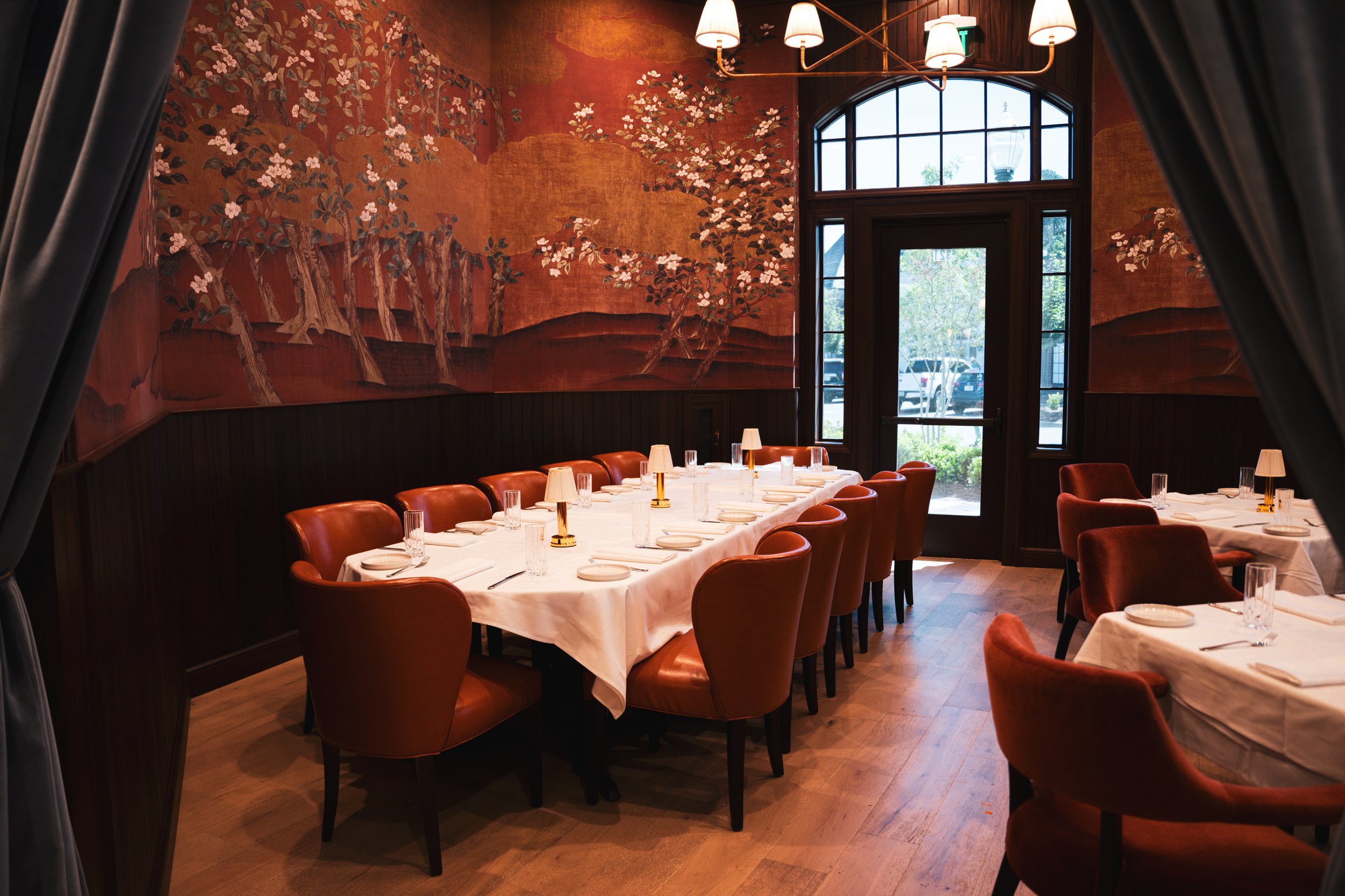 Elegant restaurant dining room with white tablecloths, brown leather chairs, small lamps, and a large mural of cherry blossoms on the wall. Large windows let in natural light.