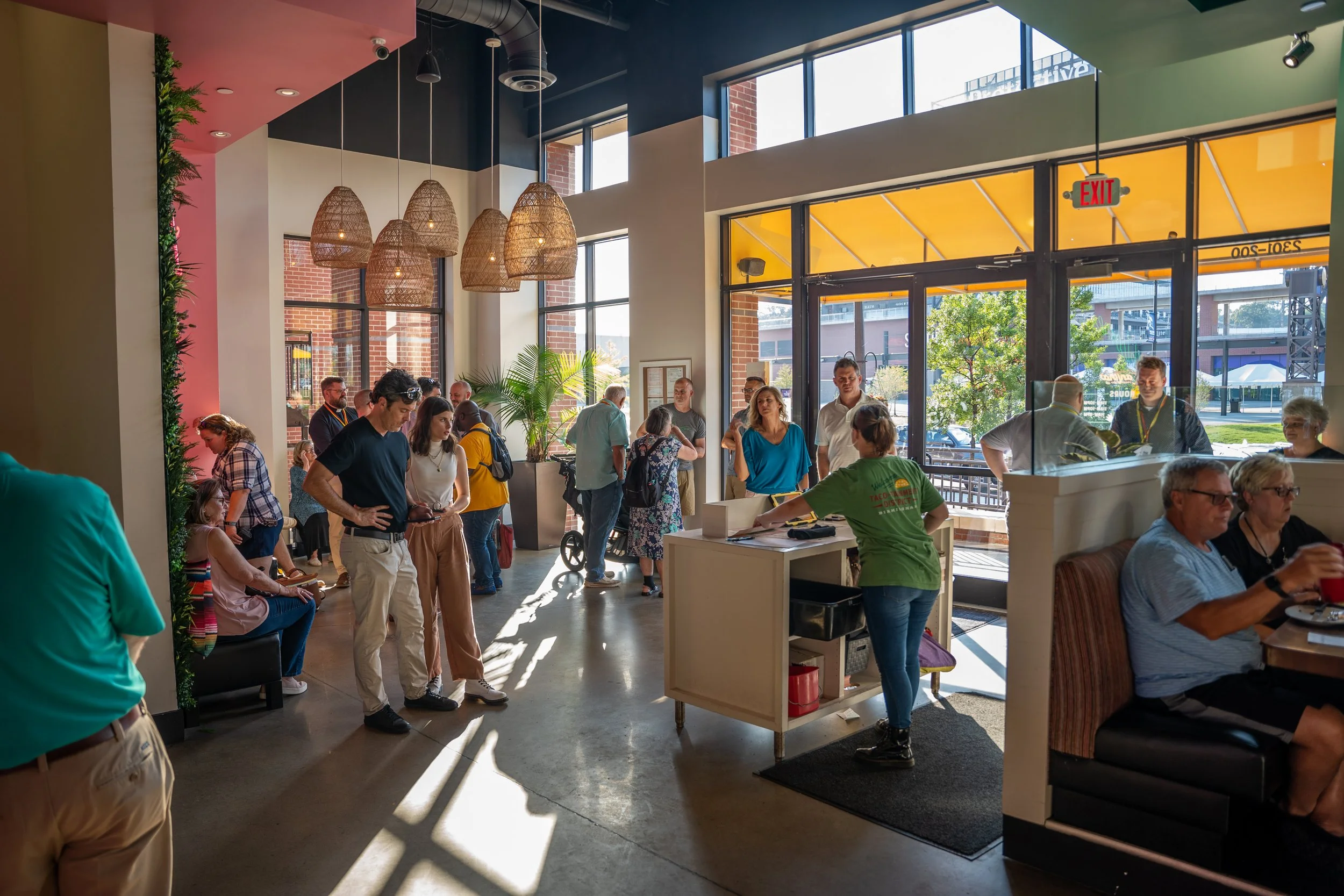 Inside a busy restaurant during daytime with customers waiting in line, being seated, and dining, featuring large windows, hanging light fixtures, and a mix of seating options.