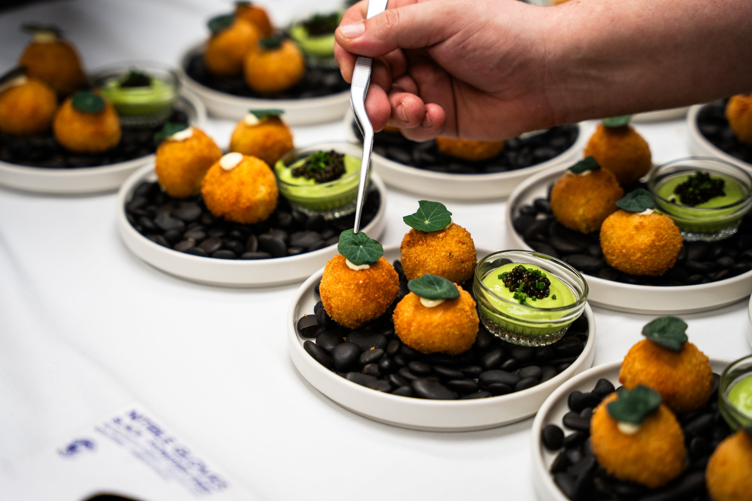 Person garnishing bite-sized appetizer with a small leaf of green herb on a white plate filled with black stones, small glass bowl of green dip or sauce, all arranged on a white table.