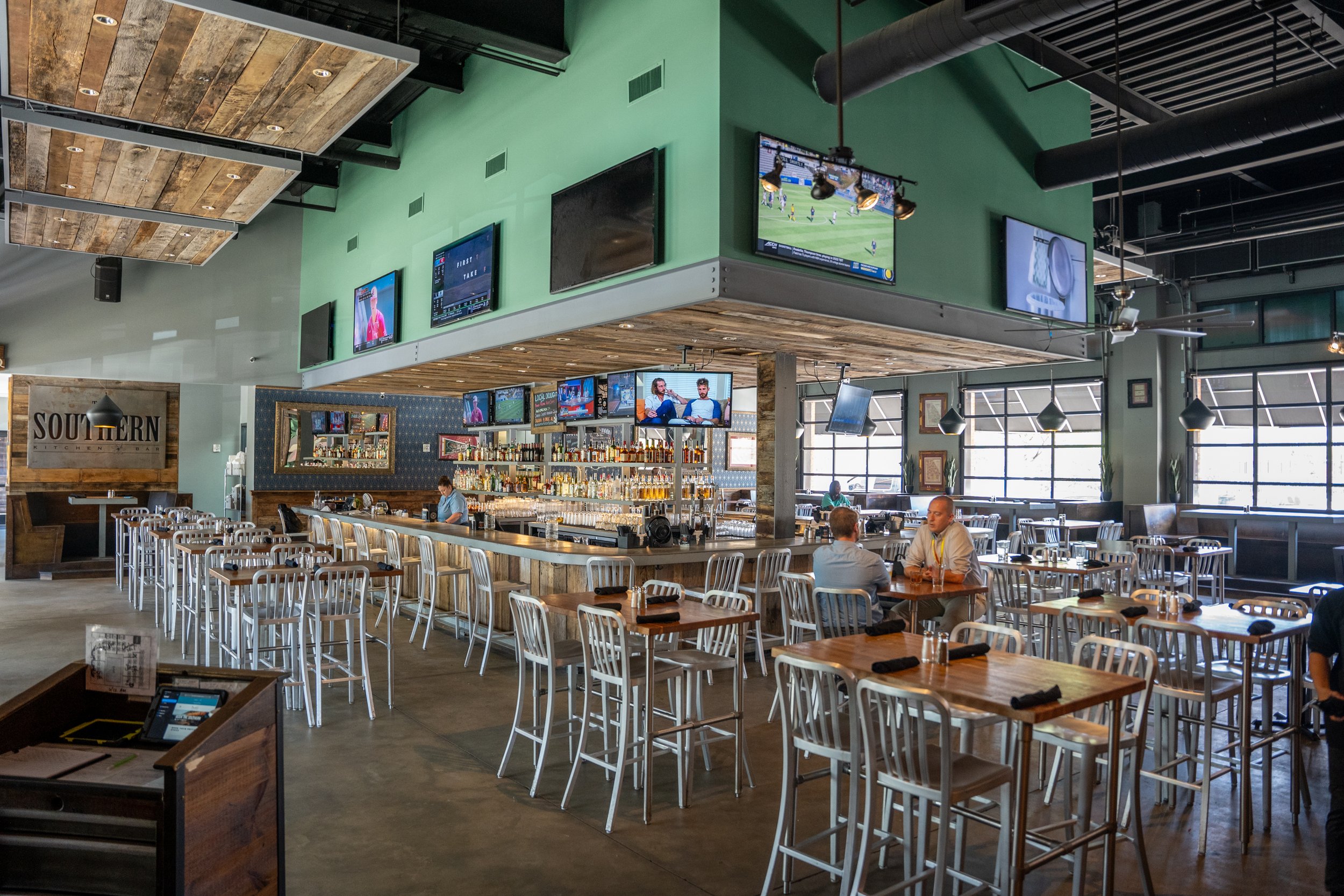 Interior of a sports bar or restaurant with high tables and chairs, multiple television screens showing sports, a bar area with bottles, and a few patrons seated and talking.