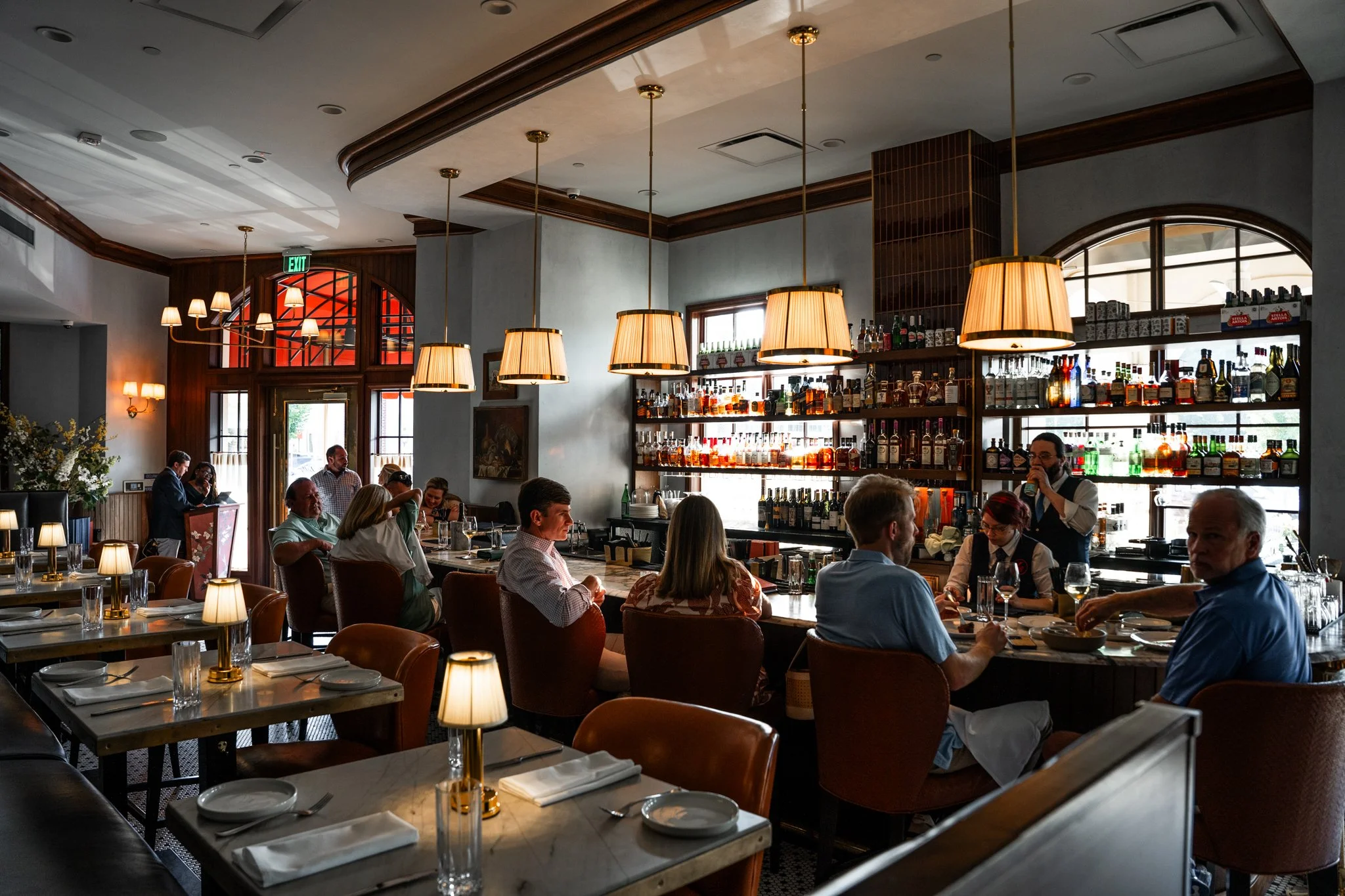 People dining at a bar counter inside a restaurant with a well-stocked liquor shelf and large windows.