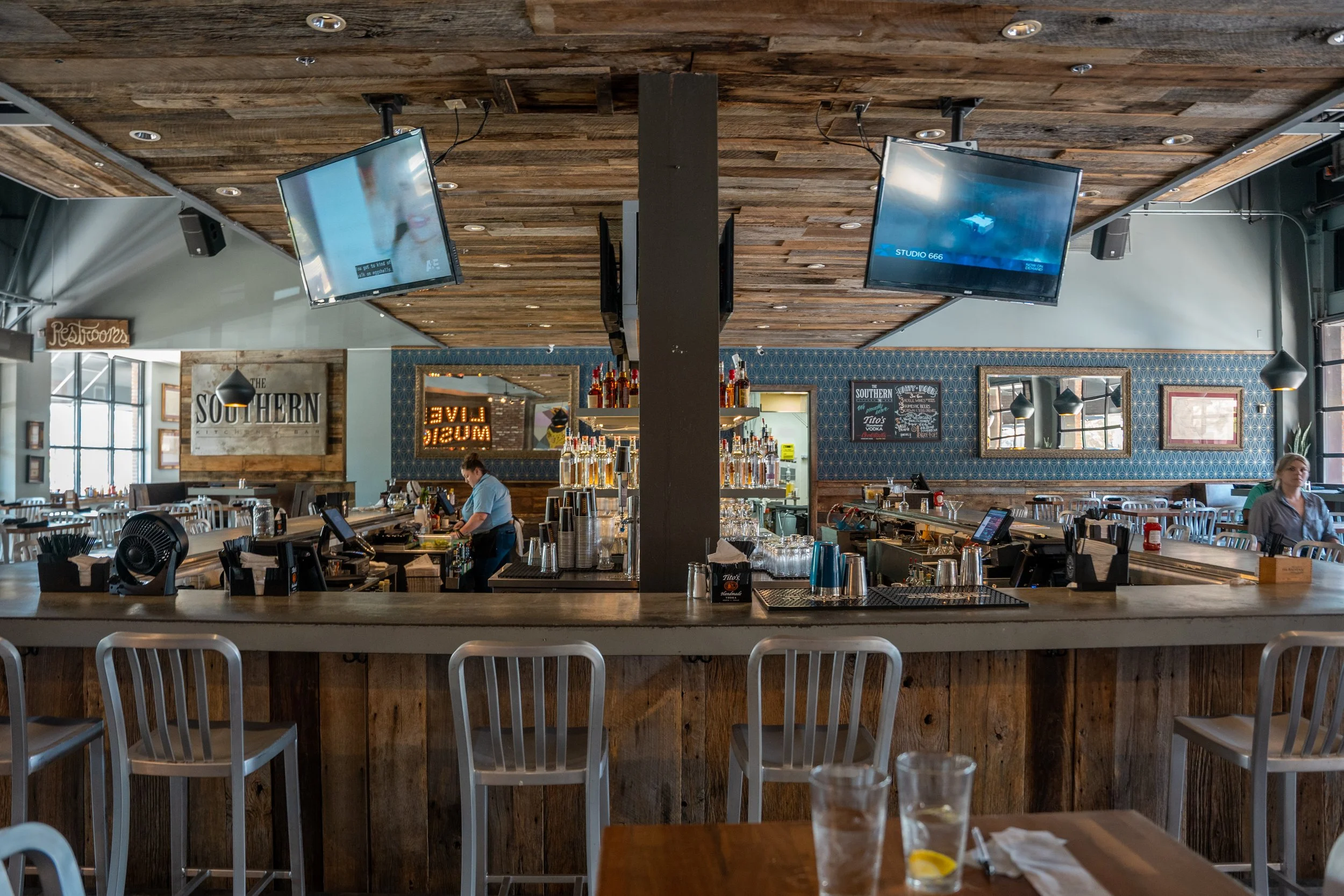 Interior of a bar and restaurant with a wooden bar counter, bar stools, television screens, and a bartender preparing drinks. There are tables and chairs on the side, with windows letting in natural light.