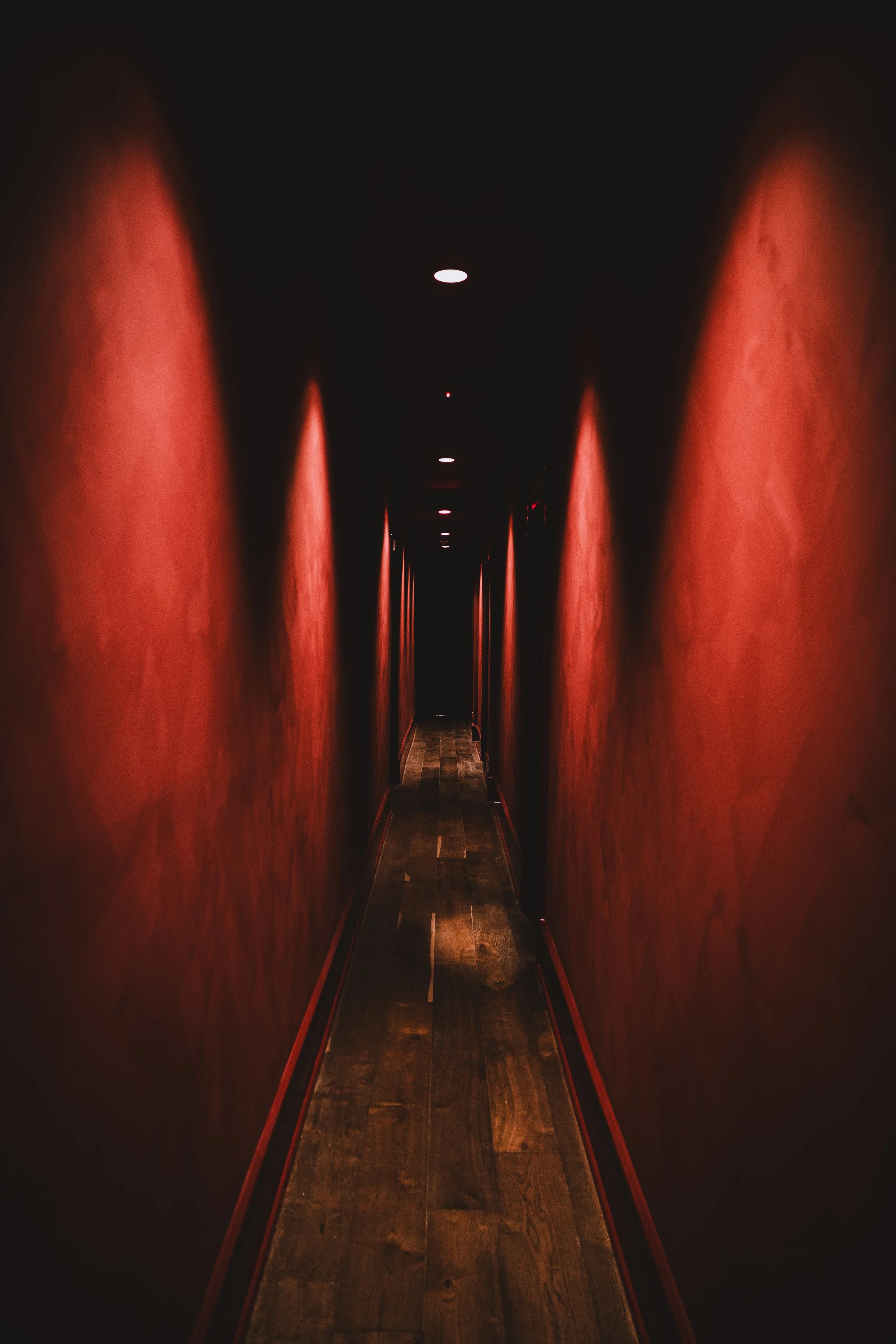Dimly lit corridor with red walls and wooden floor, illuminated by small ceiling lights.