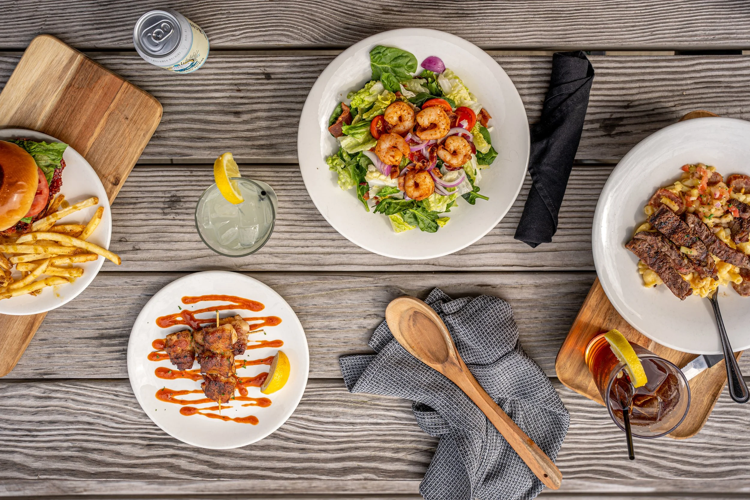 A rustic wooden table set with various dishes of food, including a salad with shrimp, a plate of French fries with a burger, a plate of grilled meat with potatoes and a glass of iced tea. There are also a couple of beverages with lemon slices and bla