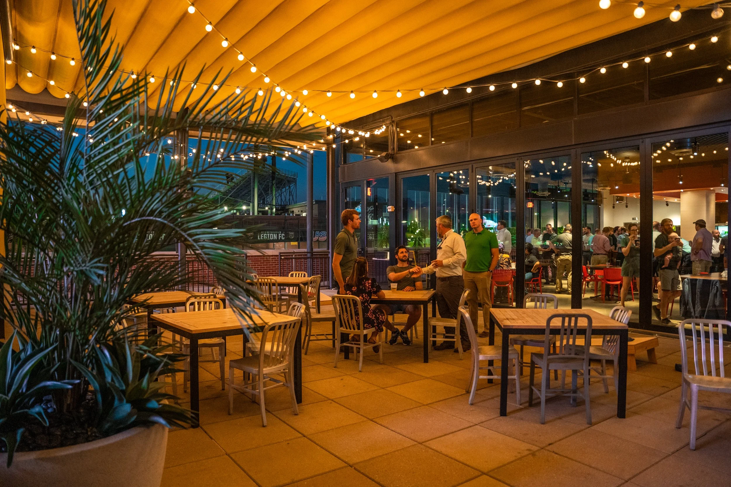 An outdoor patio of a restaurant or bar at night, with string lights overhead, wooden tables and chairs, and a large potted plant in the foreground. Inside, people are gathered, chatting and some are waiting in line at the counter.
