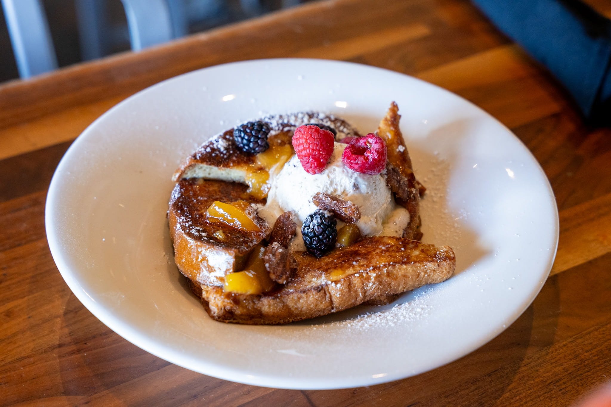 Fried French toast topped with whipped cream, fresh raspberries, blackberries, and caramelized bananas, served on a white plate with powdered sugar.