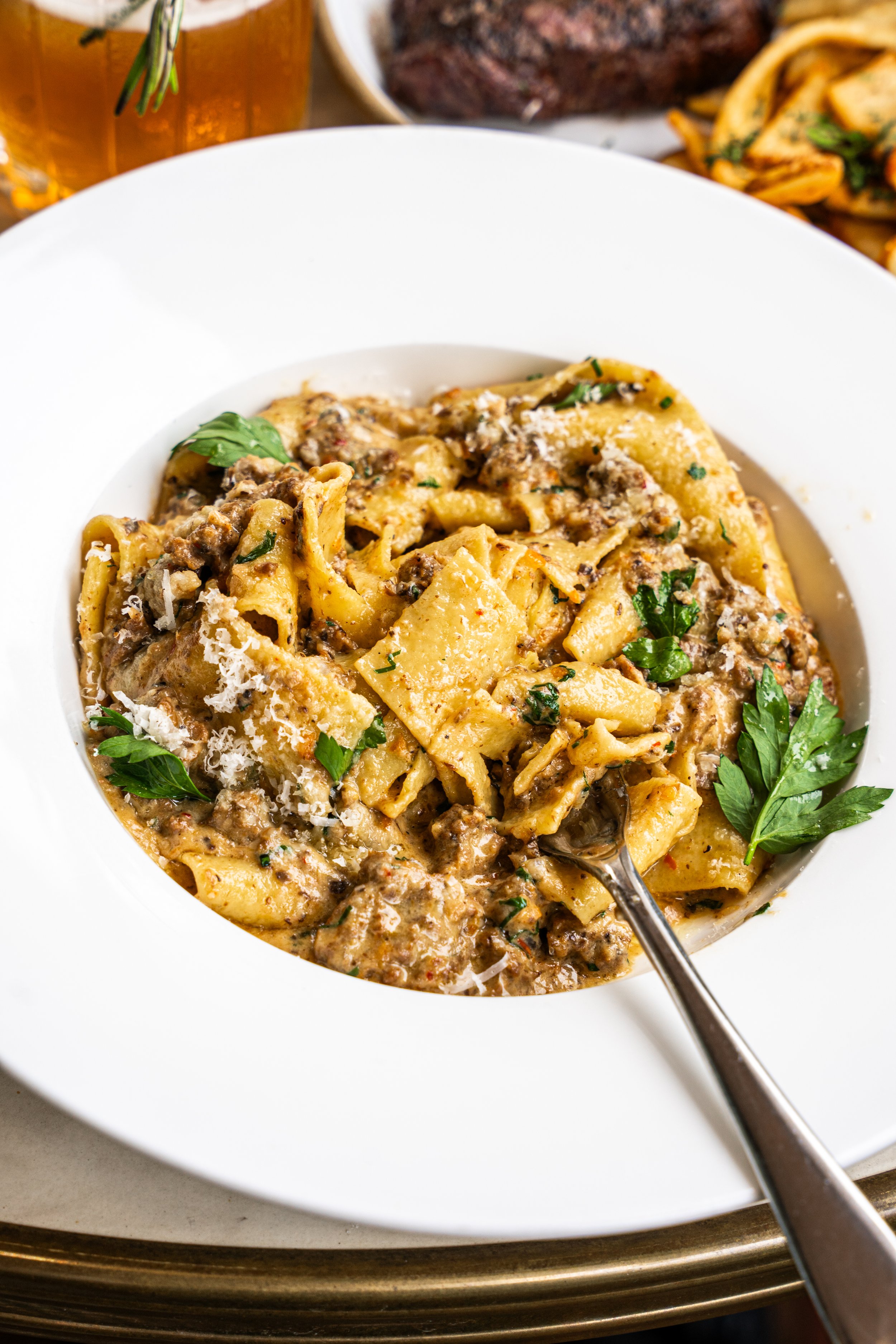 Bowl of beef and pasta with herbs, grated cheese, and a spoon in white dish on a tray.