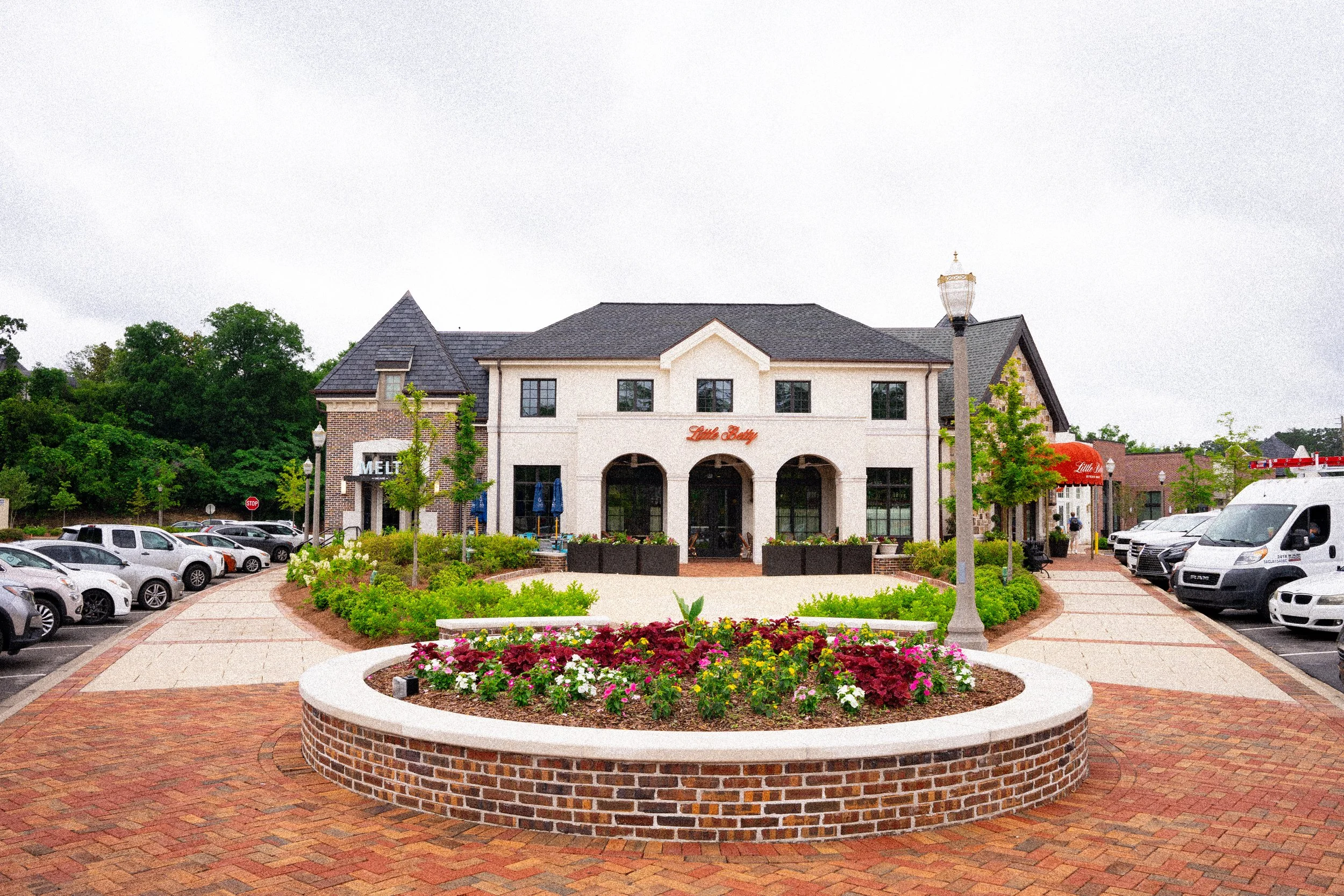 A shopping center with a flower bed in the foreground, surrounded by parked cars and trees. The building has a sign that reads 'Little Betty' above the entrance, with outdoor seating and a smaller store called 'MELT' on the left.