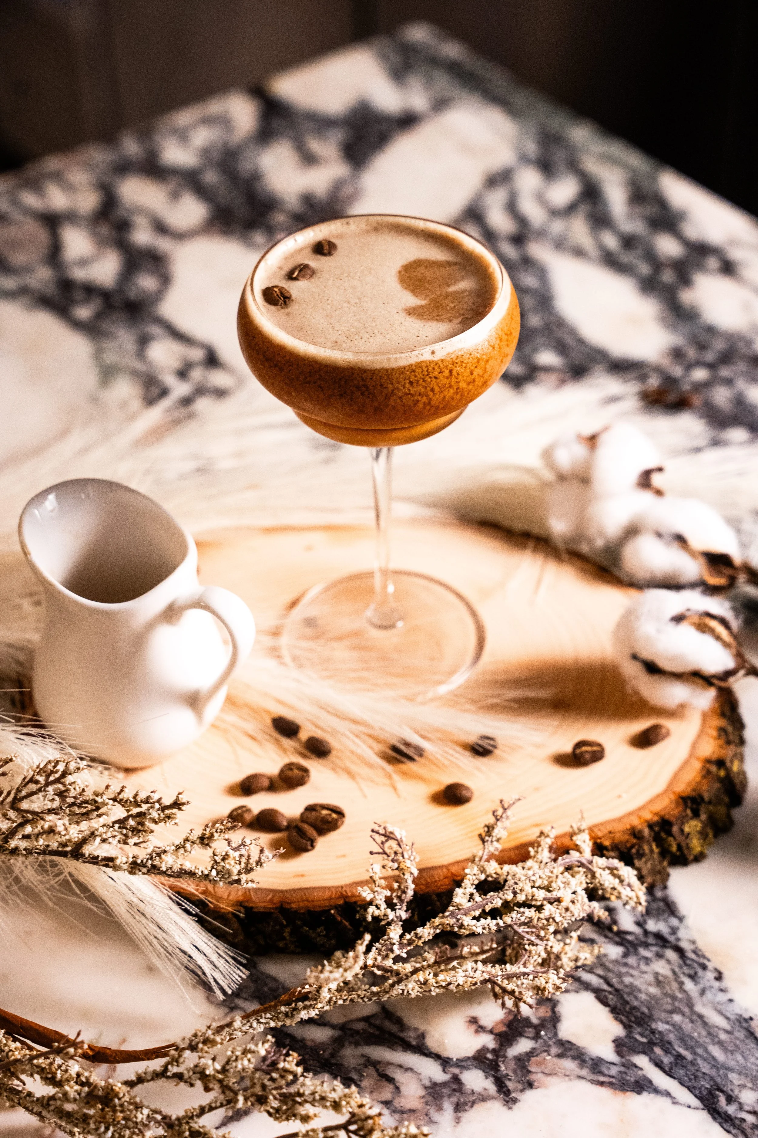 A glass of coffee with foam and three coffee beans on top, placed on a wooden slice with a small white creamer pitcher, coffee beans, and cotton plant stems around it, on a marble surface.