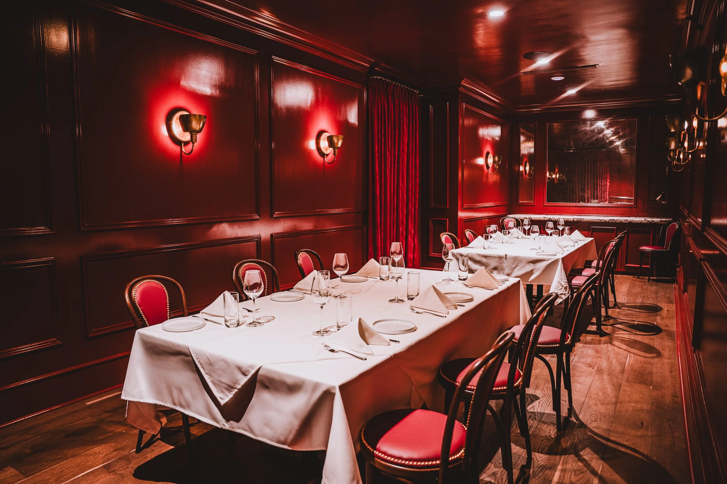 Empty restaurant dining room with white tablecloths, wine glasses, and neatly folded napkins, surrounded by red and black chairs, with red painted walls and wall sconces.