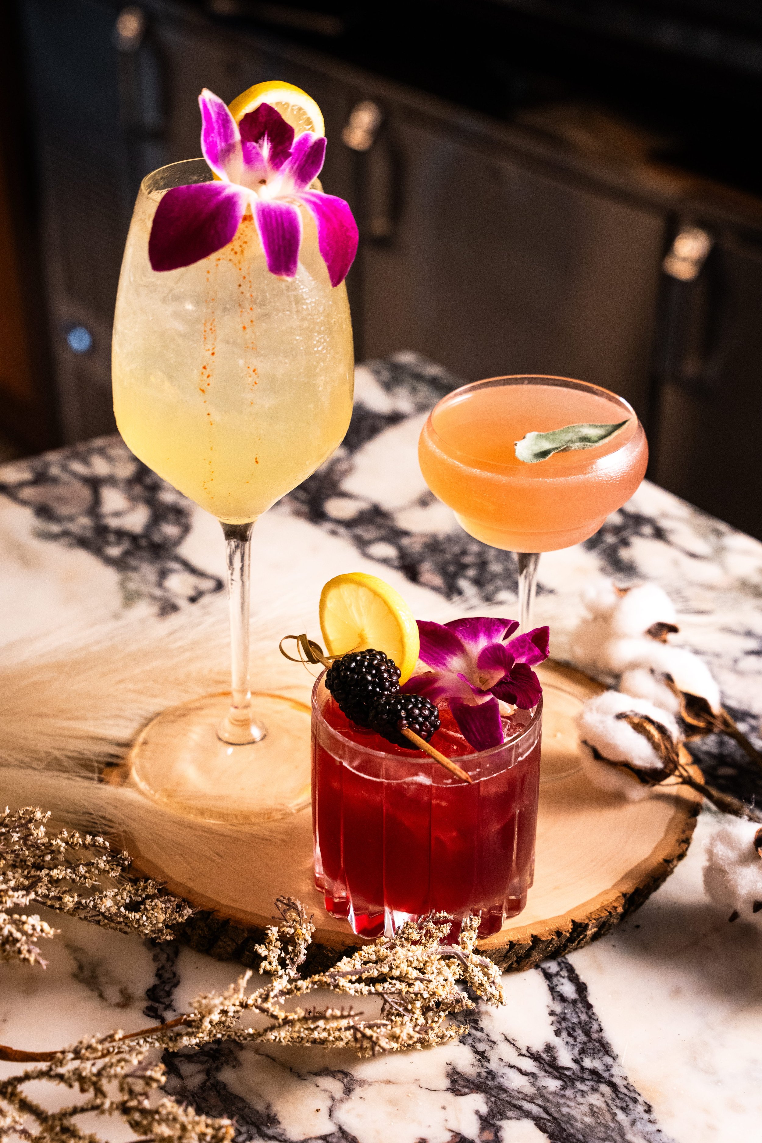 Three cocktails decorated with edible flowers, lemon slices, and berries on a wooden serving board, with cotton branches and a marble surface.