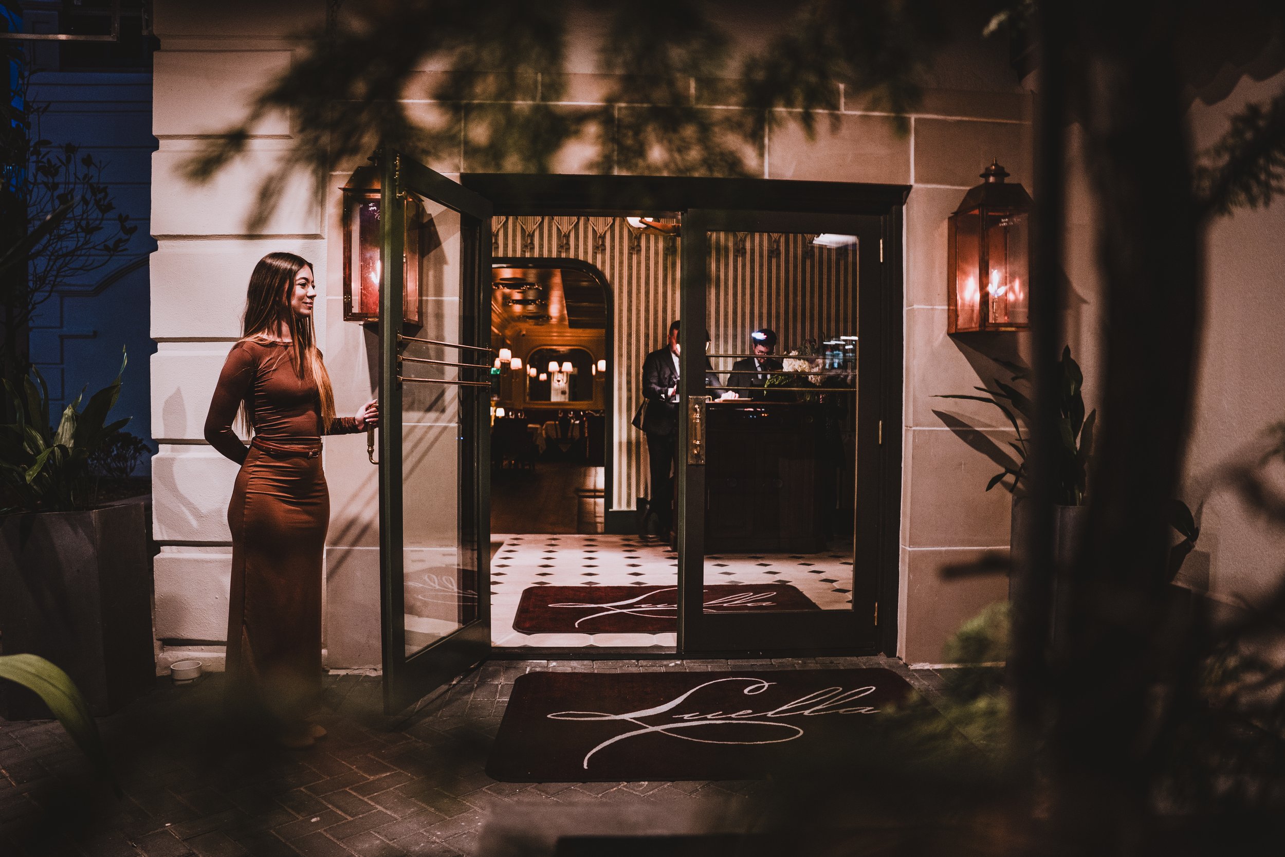 A woman in a brown dress standing outside a restaurant at night. The restaurant's entrance is open, revealing a dimly lit interior with a reception counter and a person working behind it. Two wall-mounted lamps are on either side of the entrance, and