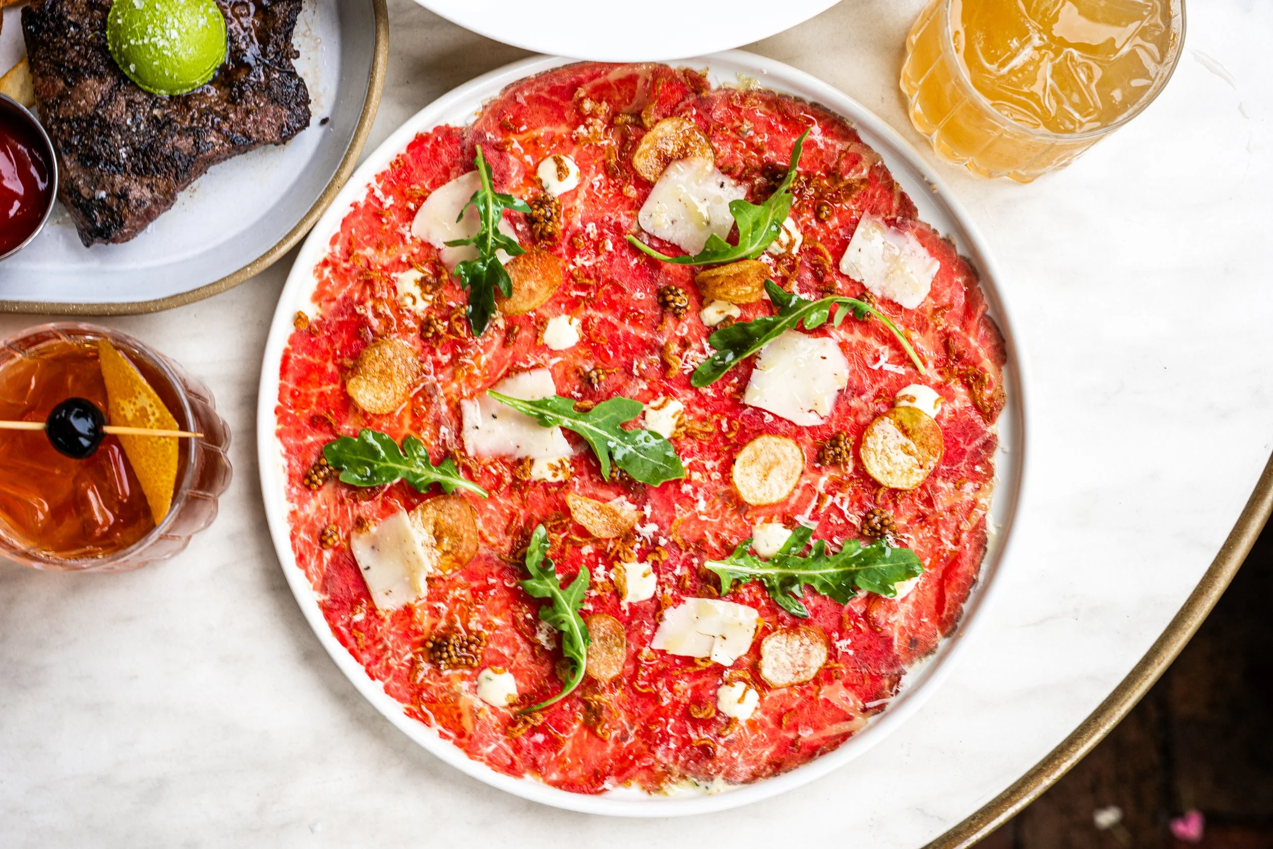 A plate of thin crust pizza with tomato sauce, topped with arugula leaves, shaved cheese, garlic chips, and mustard seeds, surrounded by glasses of iced drinks and a plate with a grilled steak with lime.