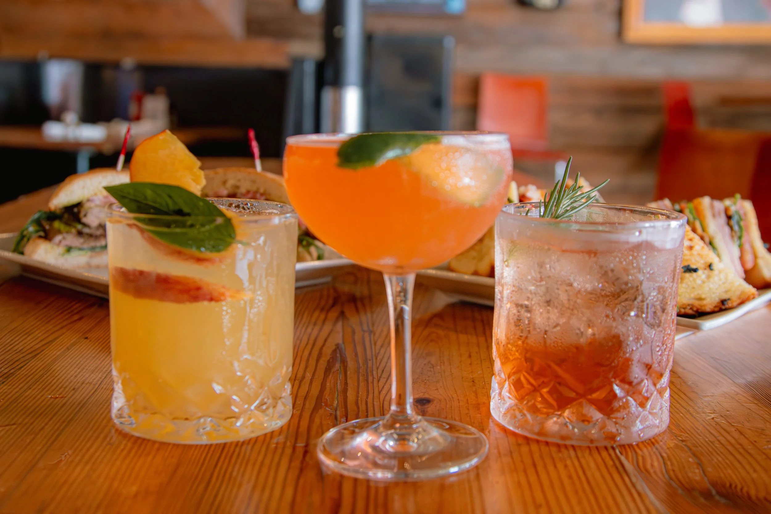 Three cocktails on a wooden table with plates of sandwiches and snacks in the background. The drinks include a yellow cocktail, an orange martini, and a pinkish cocktail, all garnished with herbs or fruit.