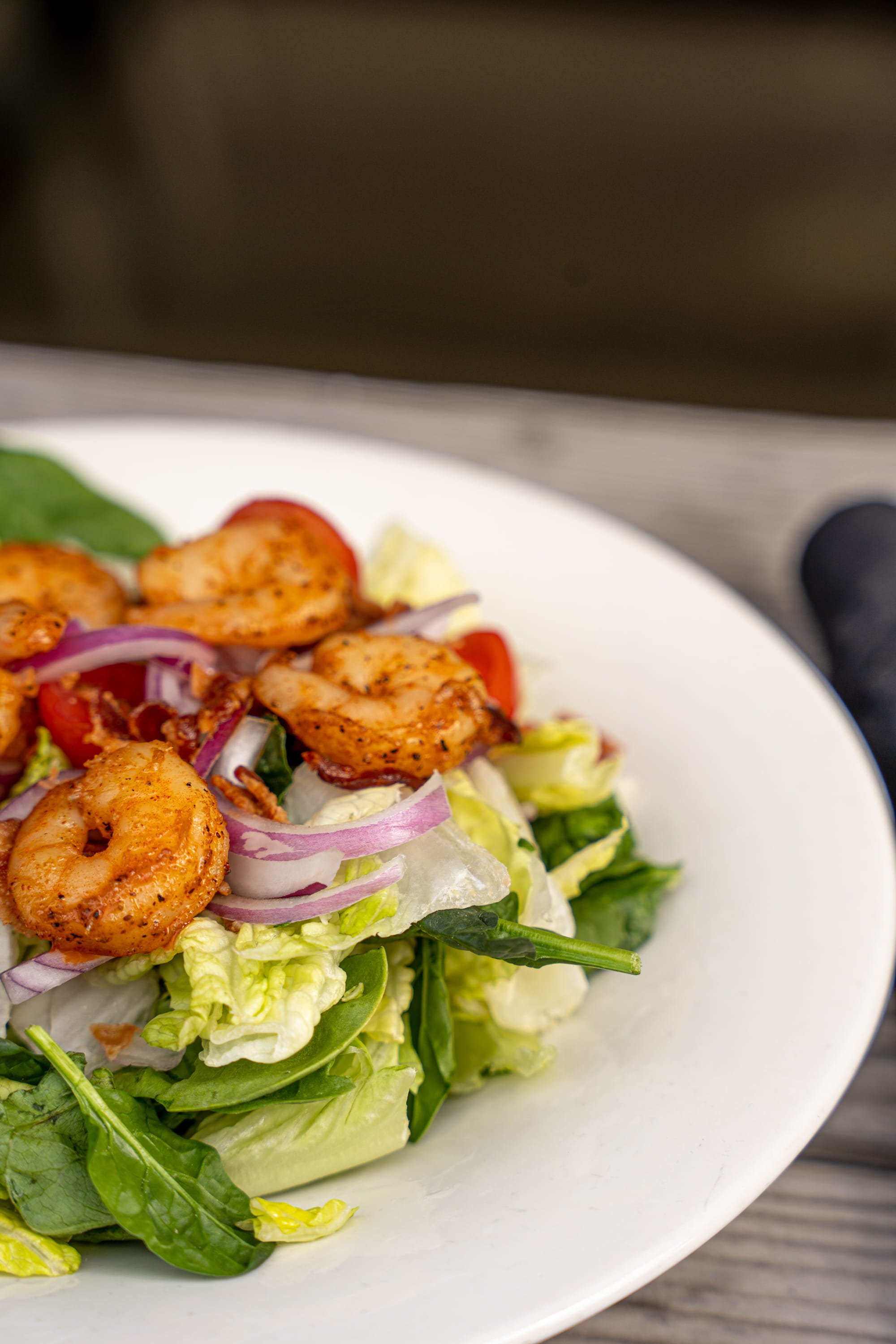 A close-up of a shrimp salad with mixed greens, cherry tomatoes, and sliced red onions on a white plate.