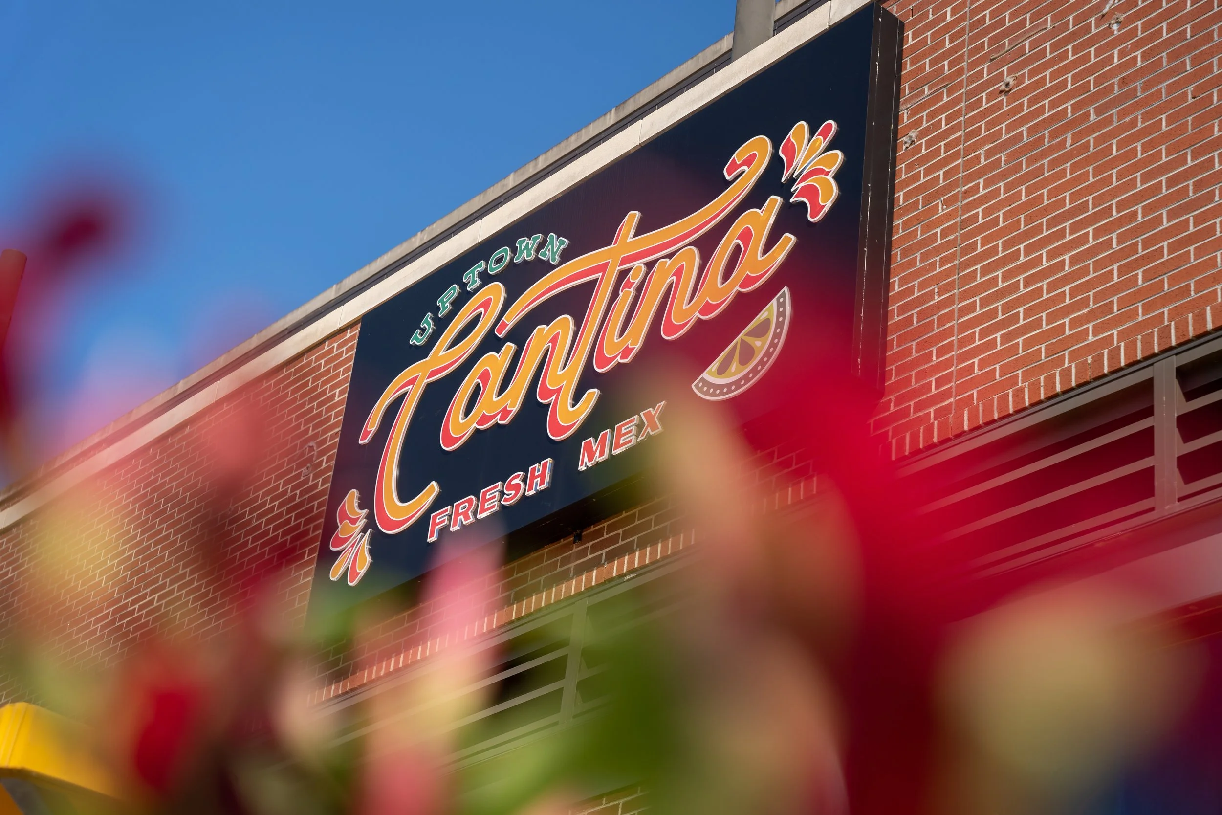 Store sign for La Pentina, a Mexican restaurant, with red brick building background, partially obscured by pink and green out-of-focus flowers in the foreground.