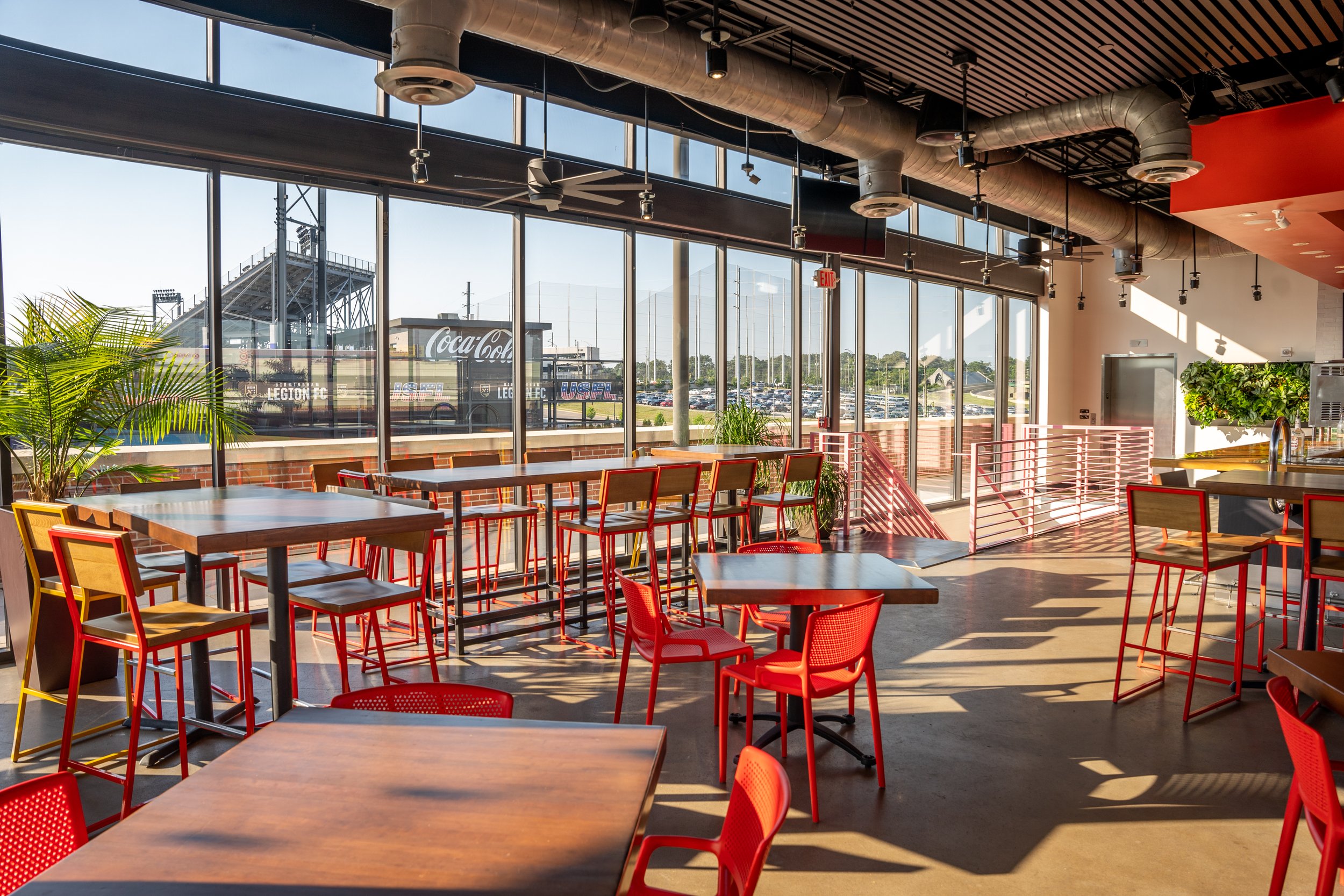Bright indoor seating area with red chairs and wooden tables, large glass windows showing an outdoor parking lot and a Coca-Cola billboard, plants, and modern decor.