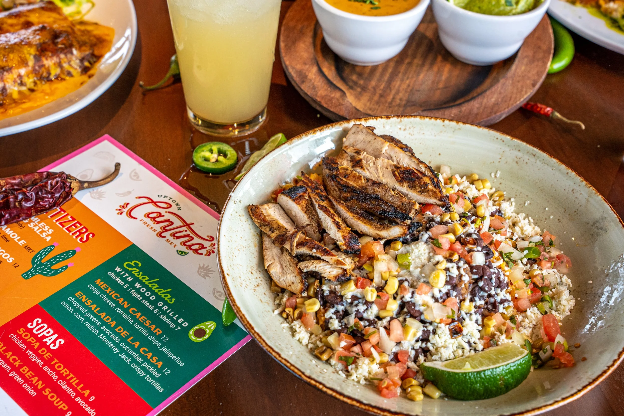 A plate of grilled chicken on top of a Mexican salad with diced vegetables, corn, beans, cheese, and crushed crackers, accompanied by a lime wedge. In the background, there is a glass of light-colored beverage, a chili pepper, a small slice of jalape