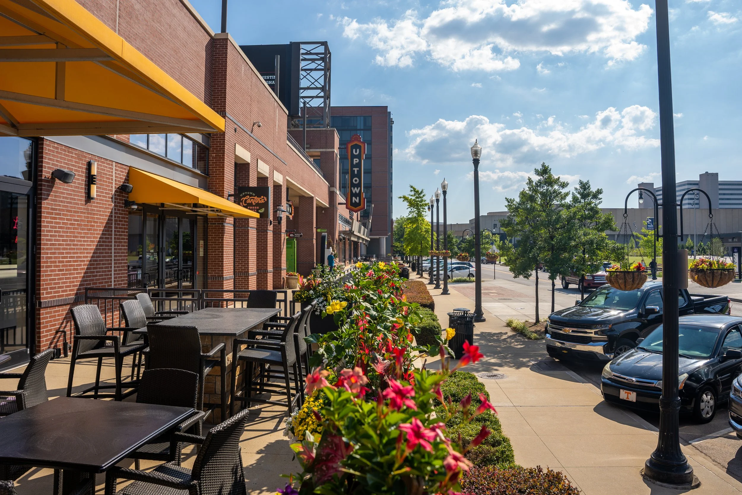 Outdoor seating area with tables and chairs outside a brick building with yellow awnings, along a sidewalk with flower planters, trees, street lamps, parked cars, and a sign reading 'Uptown' in the background on a sunny day.
