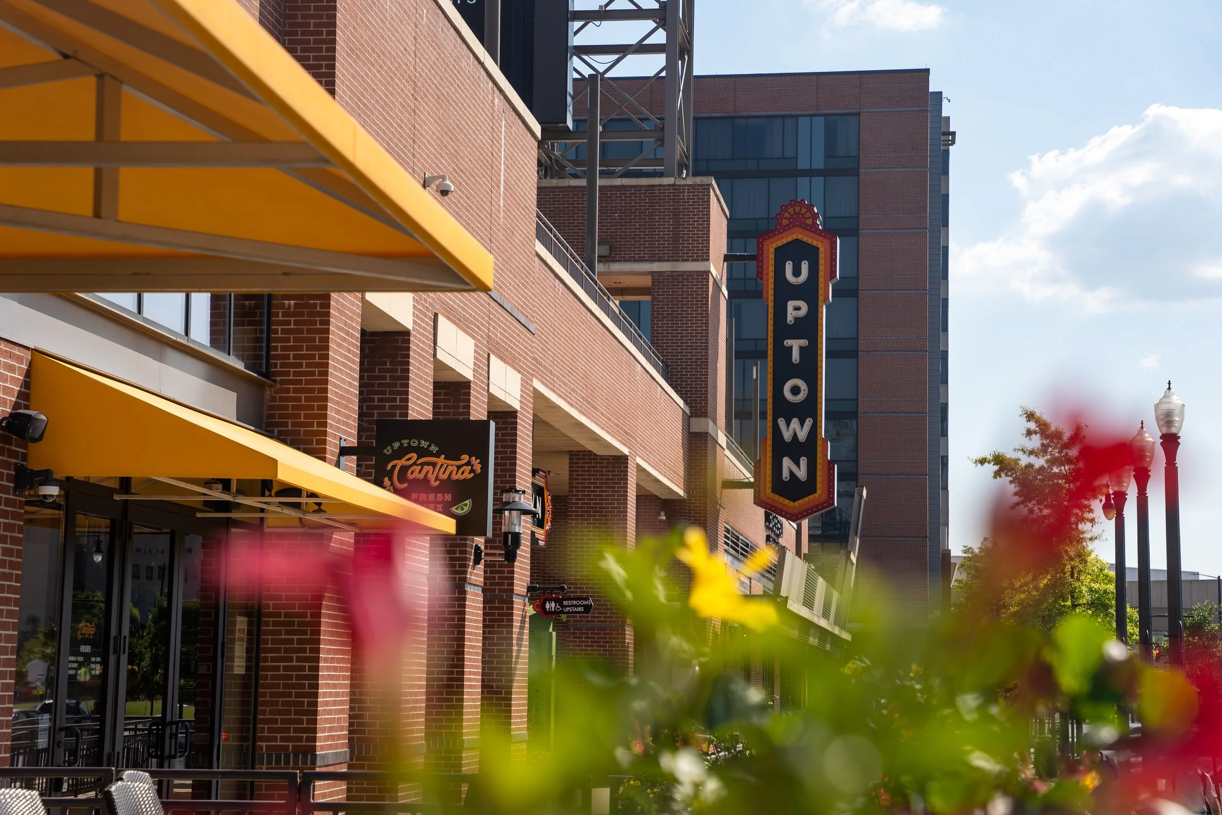 Street view of Uptown area with brick buildings, yellow awnings, a vertical Uptown sign, outdoor seating, and blurred pink and red flowers in foreground. Clear sky with some clouds.