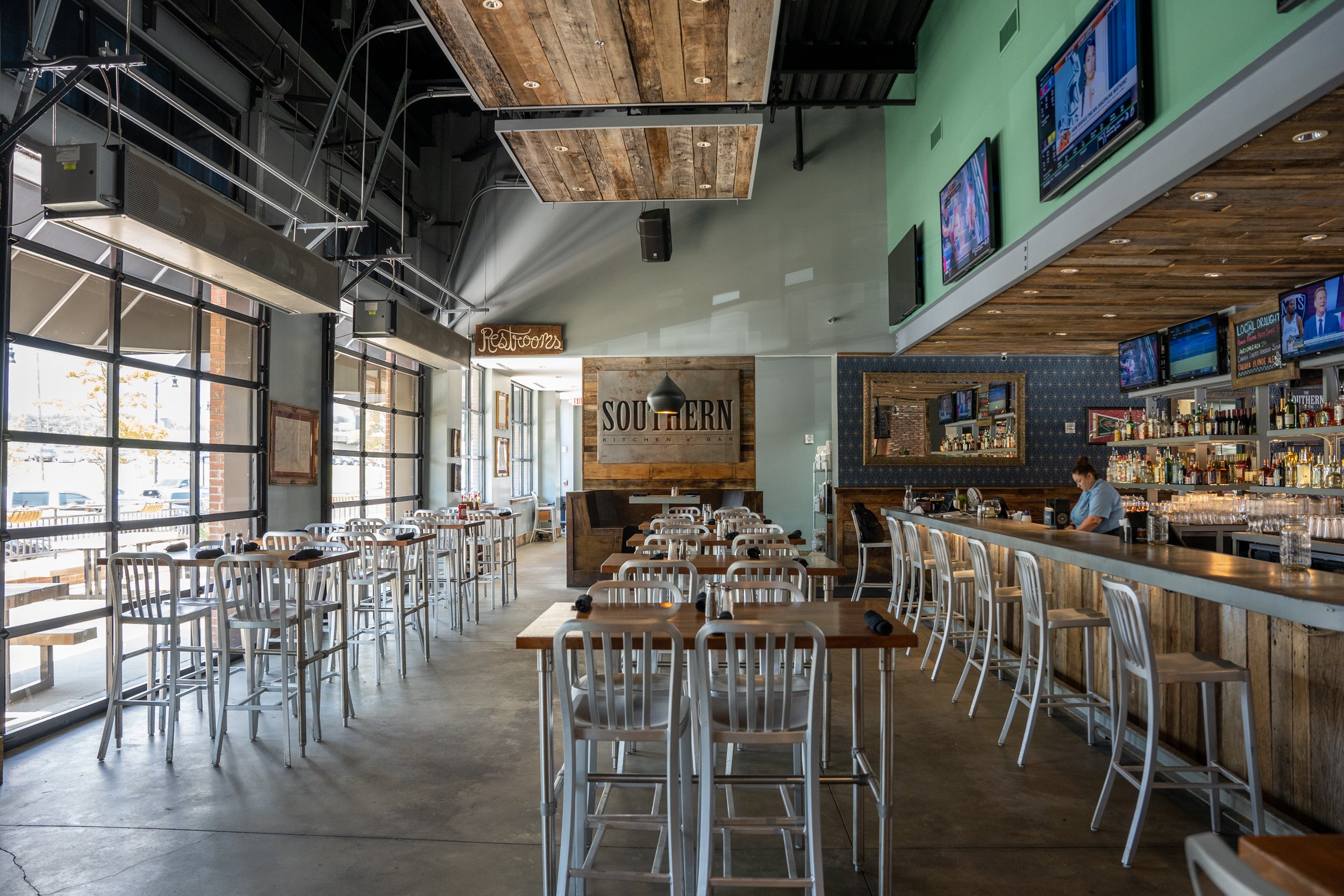 Interior of a modern restaurant or bar with high tables, bar counter, and multiple television screens on the wall, with a bartender preparing drinks and a sign reading 'Southern.'