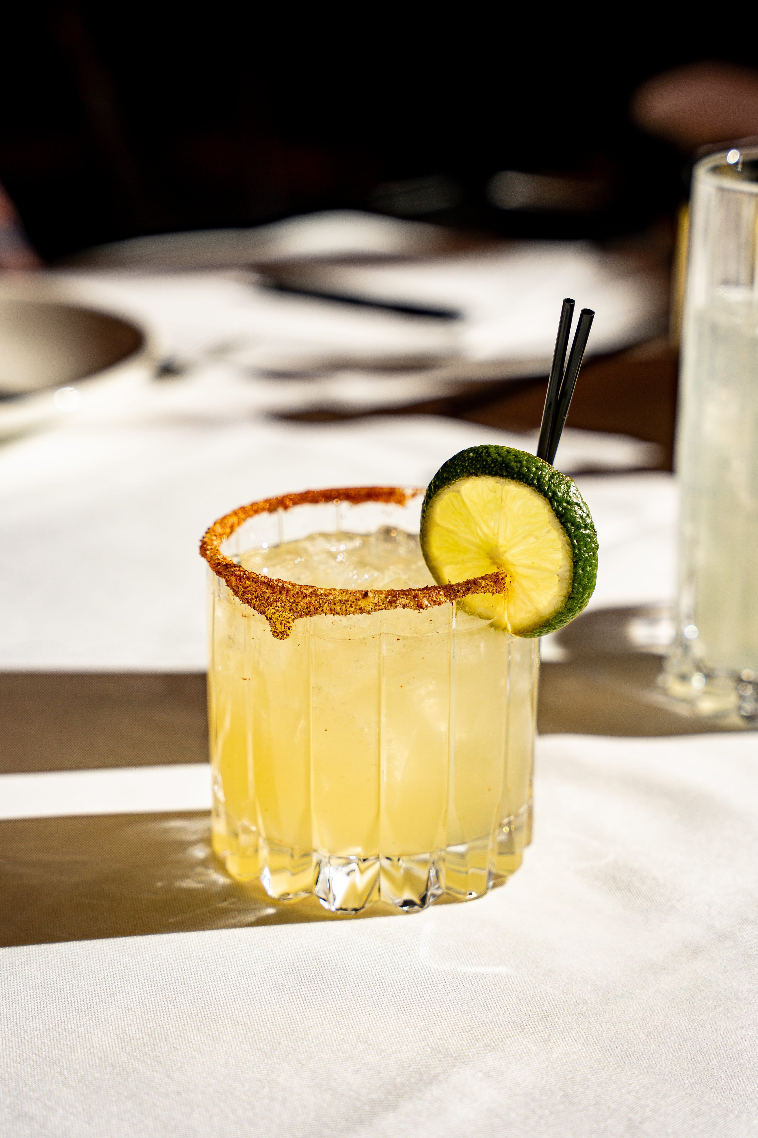 A yellow cocktail in a clear glass with a salted rim, garnished with a slice of lime, and two black straws, on a white tablecloth.