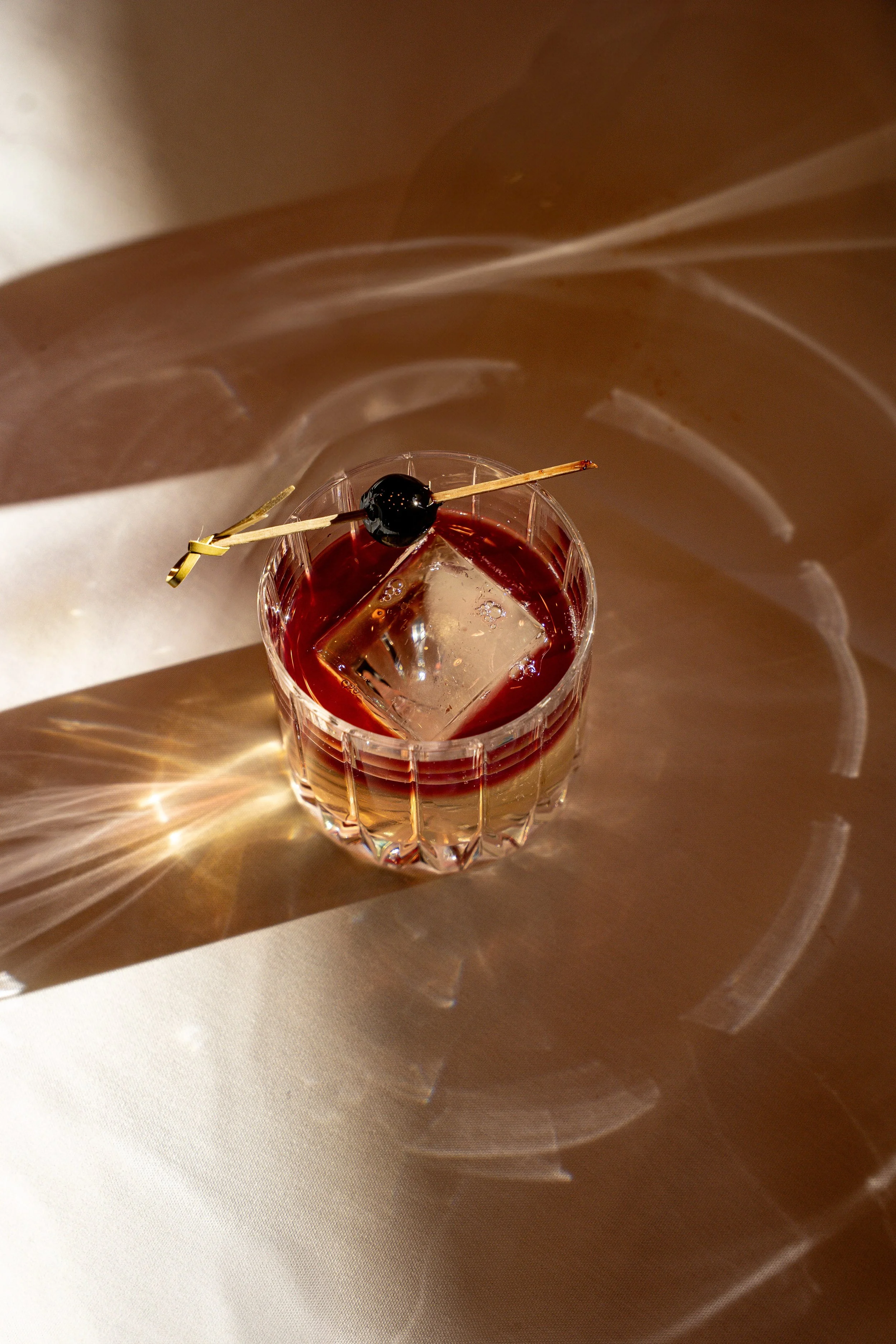 A glass of red cocktail with a large ice cube and a black cherry garnish, placed on a reflective surface with distorted light patterns.