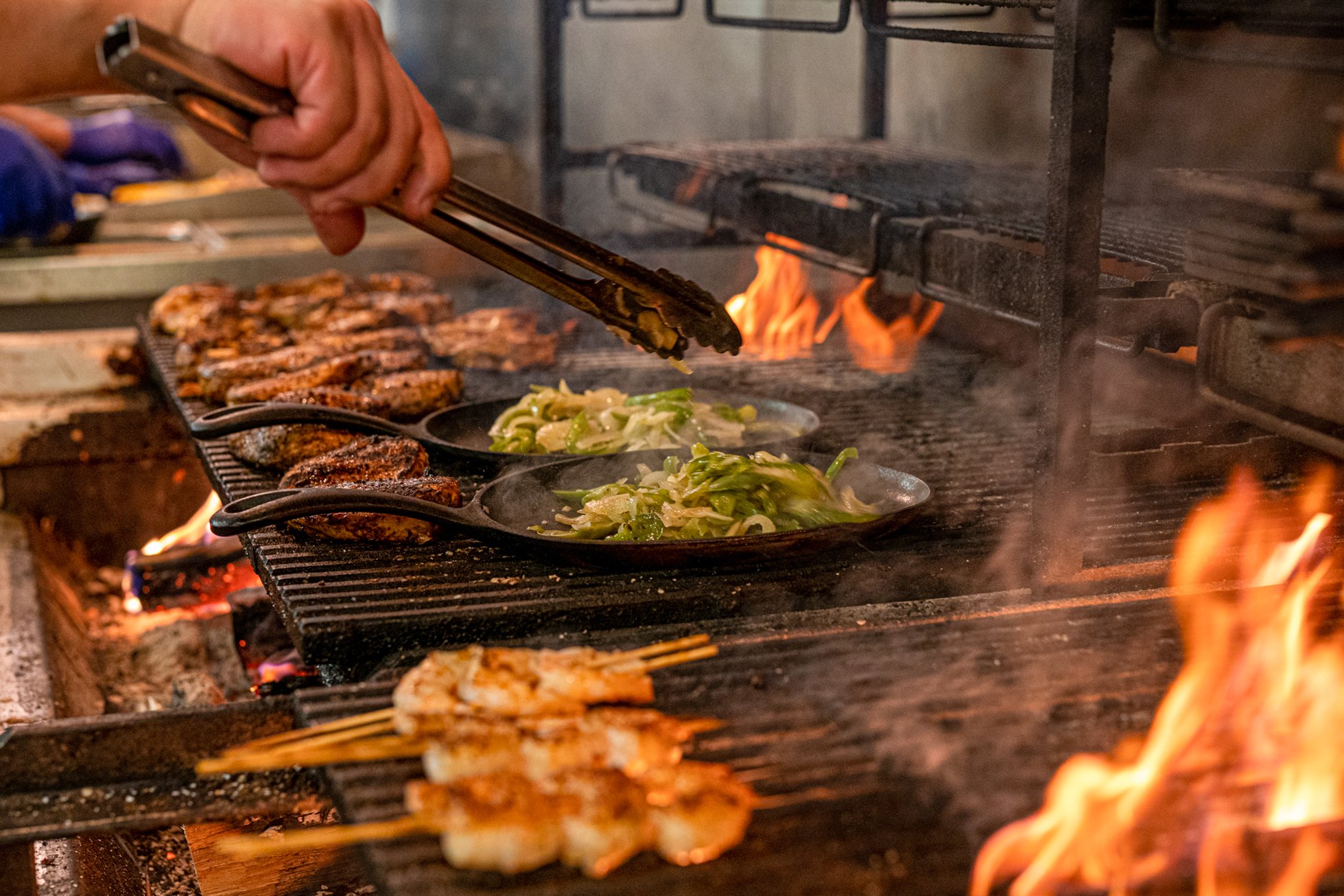 A chef cooks meat and vegetables on a grill with open flames, using tongs to handle the food.