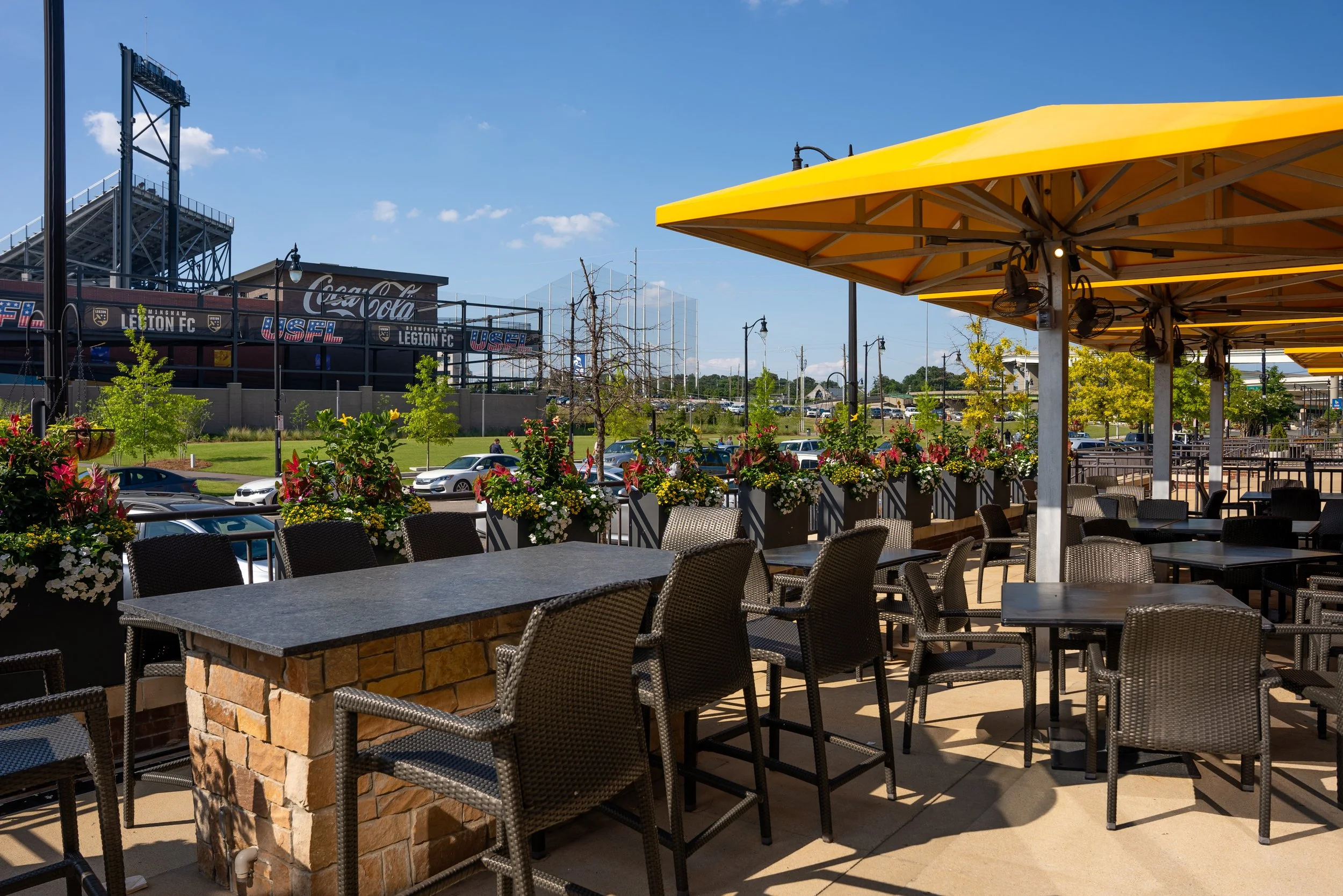 Outdoor restaurant seating area with tables, chairs, and yellow umbrellas, overlooking a parking lot with trees and a sports stadium in the background.