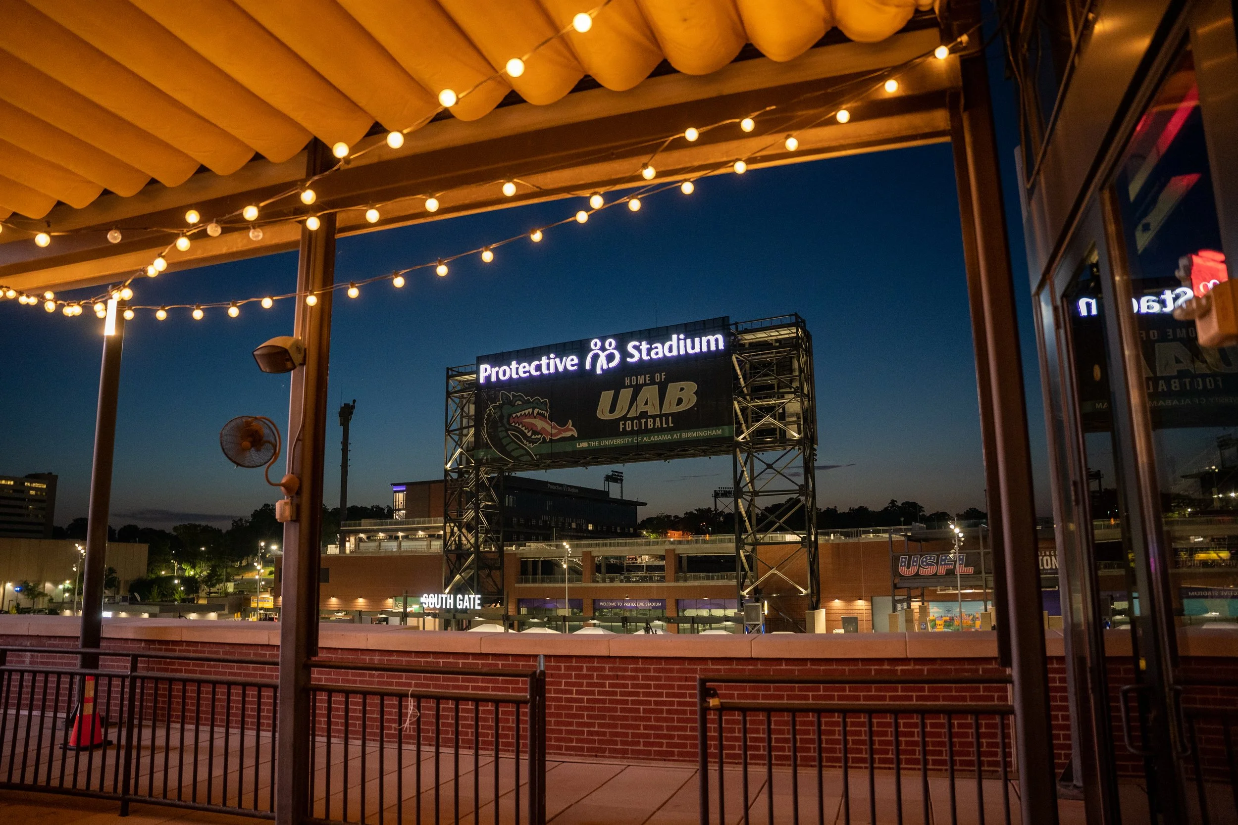 View of a football stadium sign at dusk, with string lights overhead, brick wall, and glass doors in the foreground.