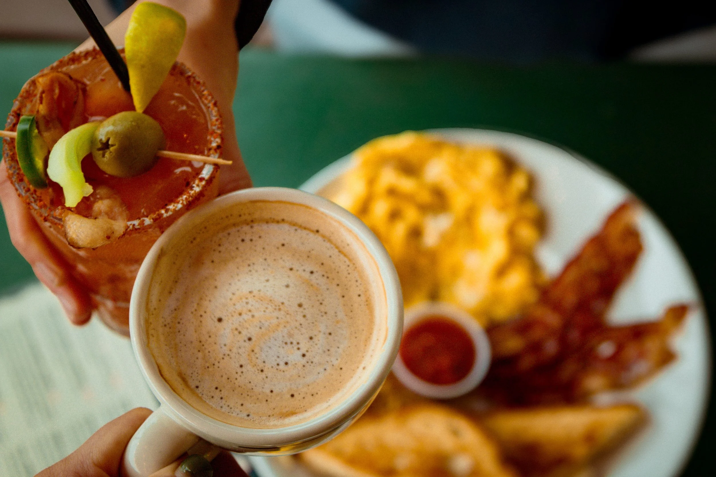 Person holding a cocktail with garnishes, a cup of coffee or latte, and a plate of breakfast food including scrambled eggs, bacon, and a small side of ketchup.