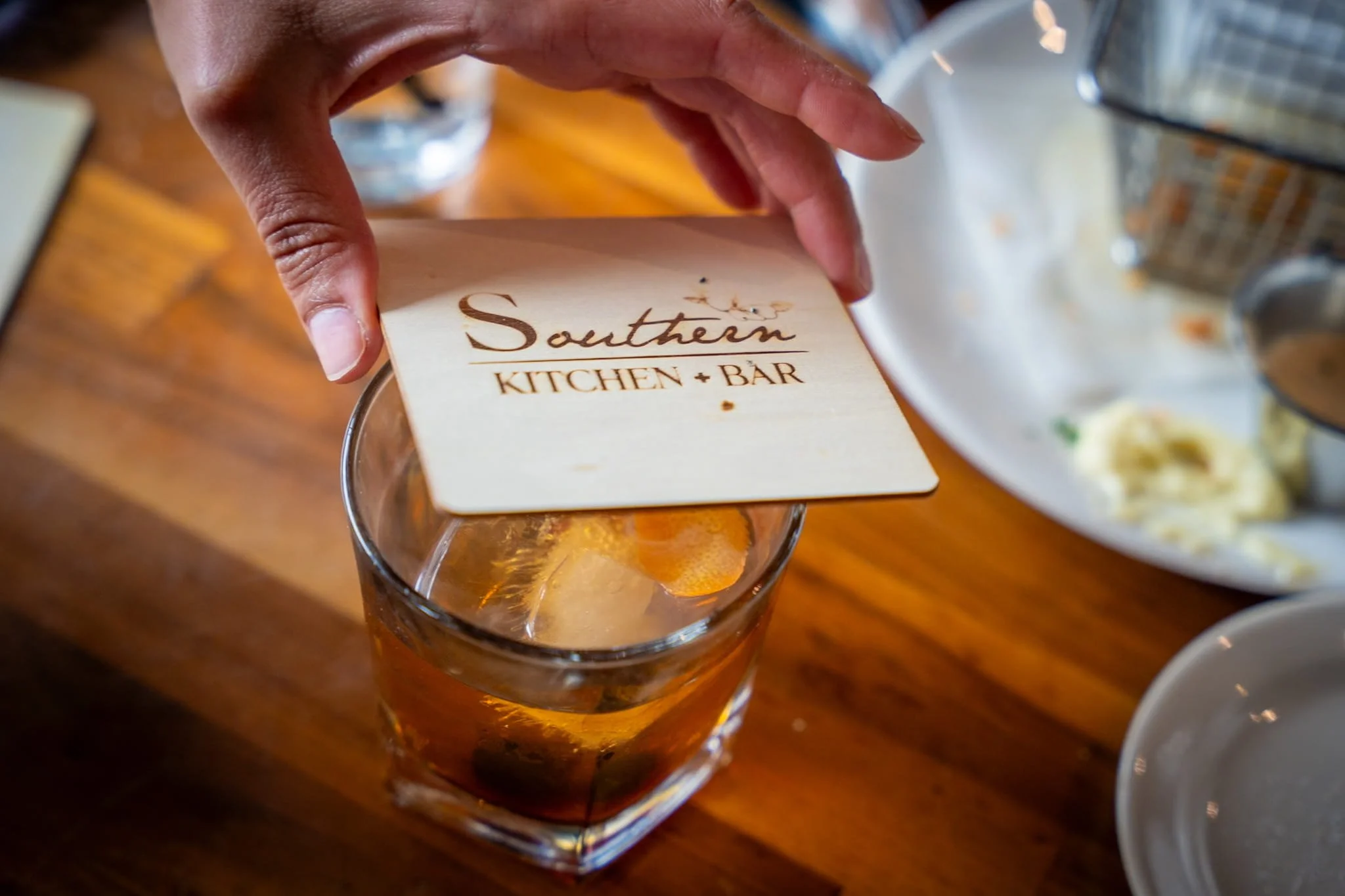 Person placing a coaster with Southern Kitchen + Bar logo on a glass of iced tea or soda on a wooden table in a restaurant.