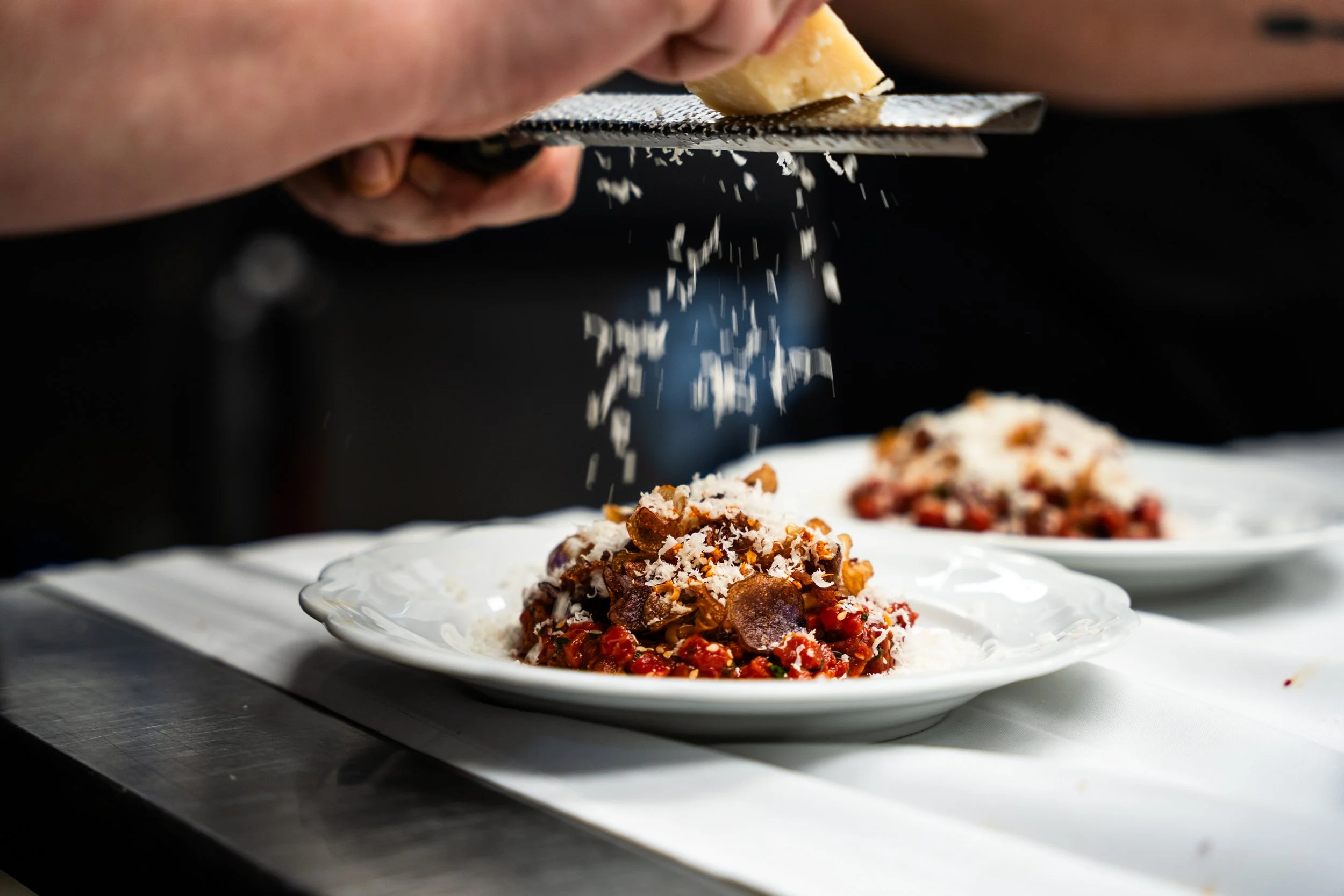 A chef grating cheese over a plated dish of meat and vegetables, with an additional identical dish in the background.