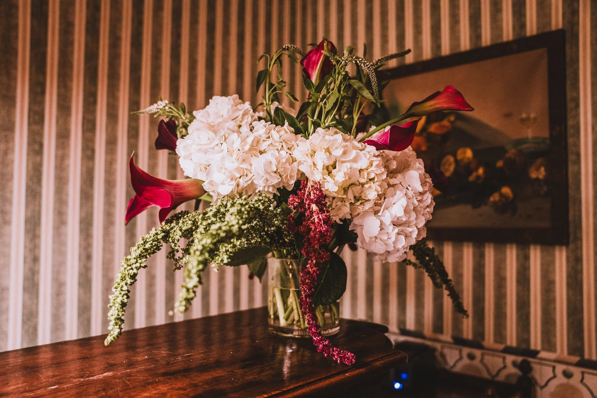 A flower arrangement in a glass vase on a wooden surface, featuring white hydrangeas, red calla lilies, and various greenery, with a mirror and striped wall in the background.