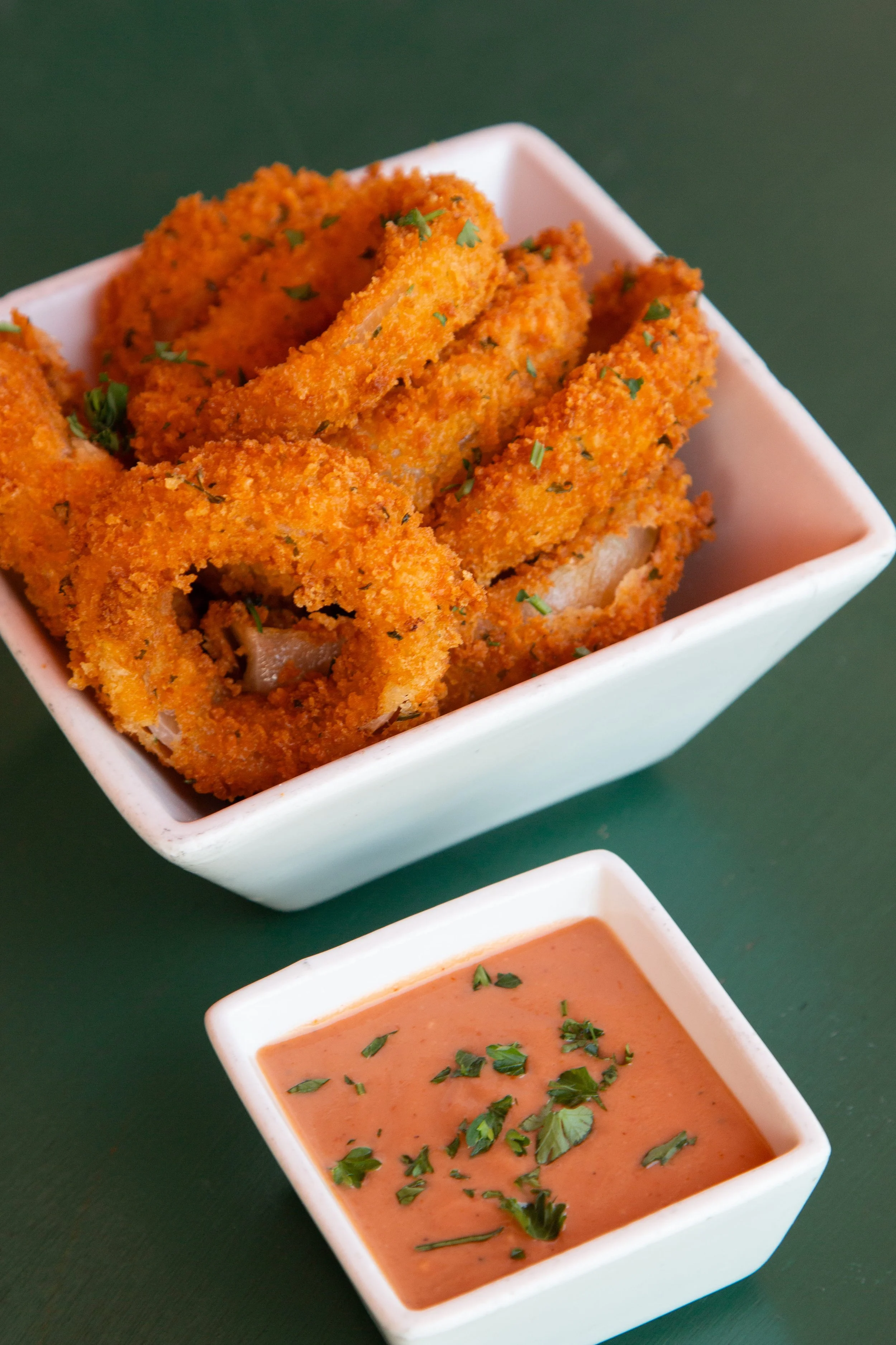 Fried onion rings garnished with herbs served with a dipping sauce in a white bowl.