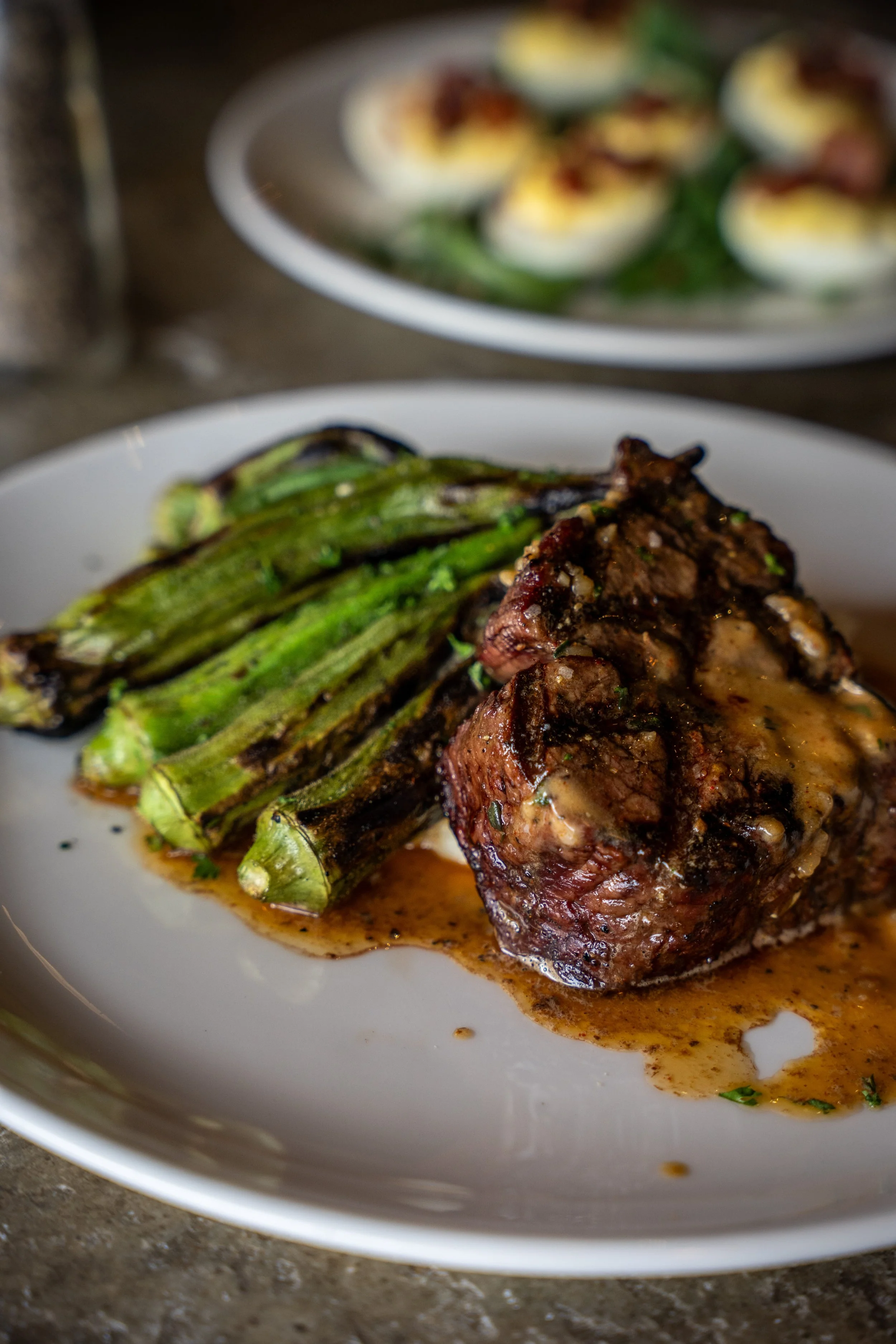 Close-up of a plated beef steak topped with gravy and grilled asparagus on a white plate with another dish of deviled eggs in the background.