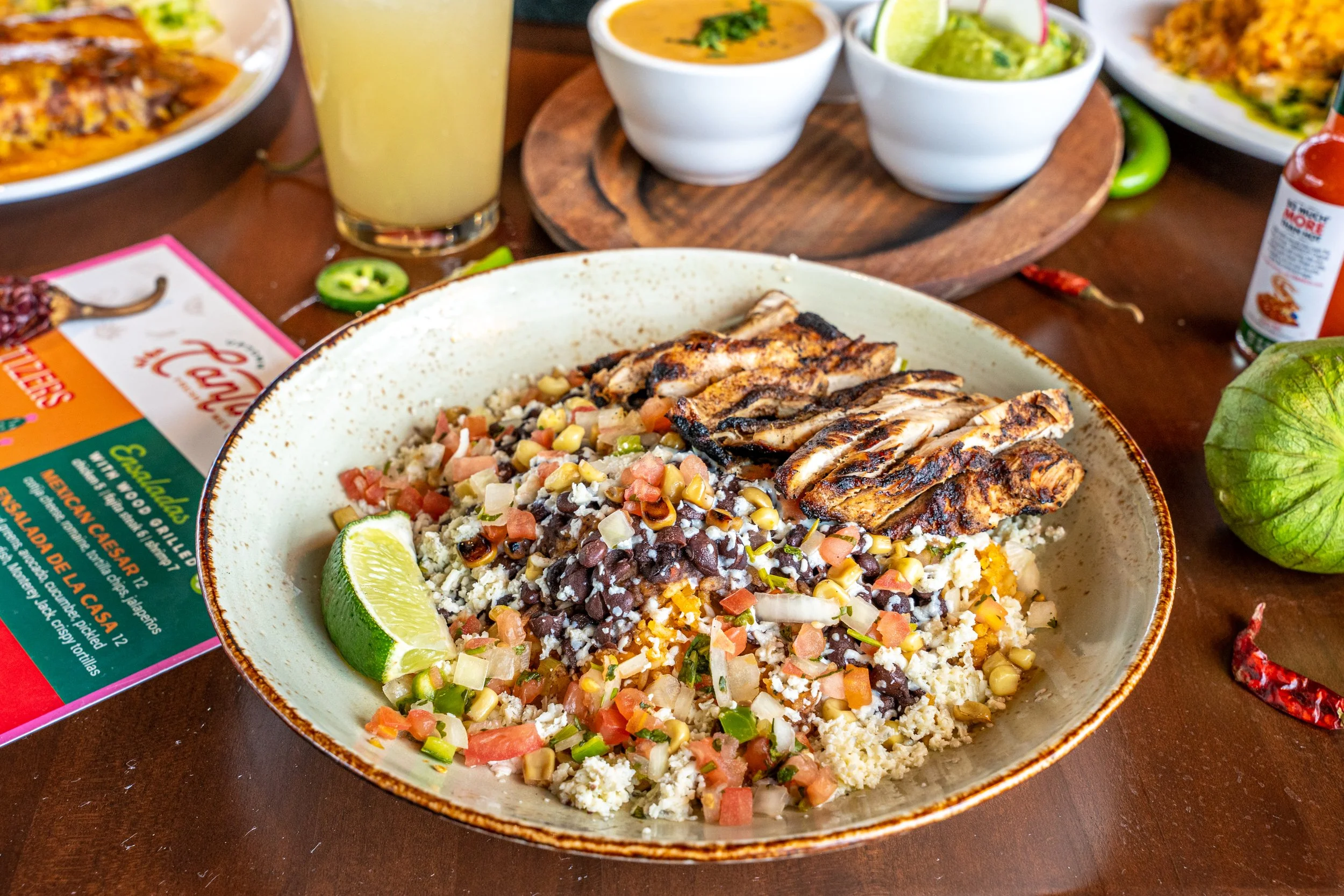 Mexican dish with grilled chicken, rice, beans, corn, chopped vegetables, lime wedge, and various side dishes on a wooden table.