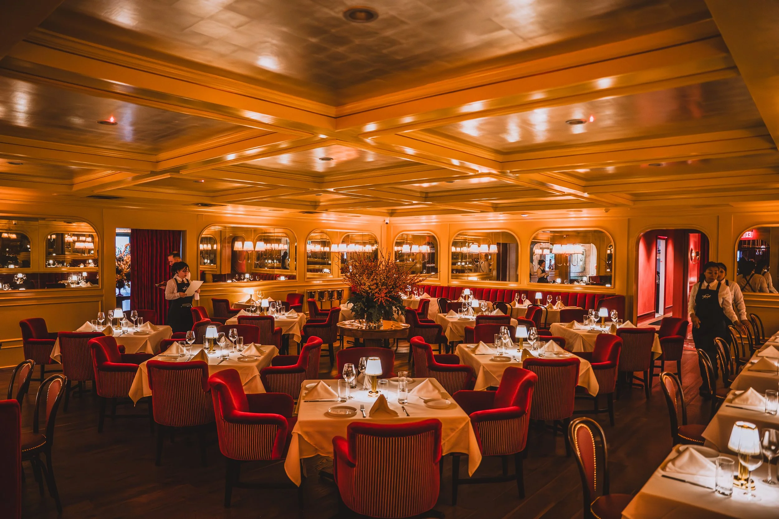 Elegant restaurant interior with multiple round tables covered with white tablecloths, red upholstered chairs, and a large floral centerpiece in the middle. Warm lighting, mirrors on the walls, and waitstaff present.