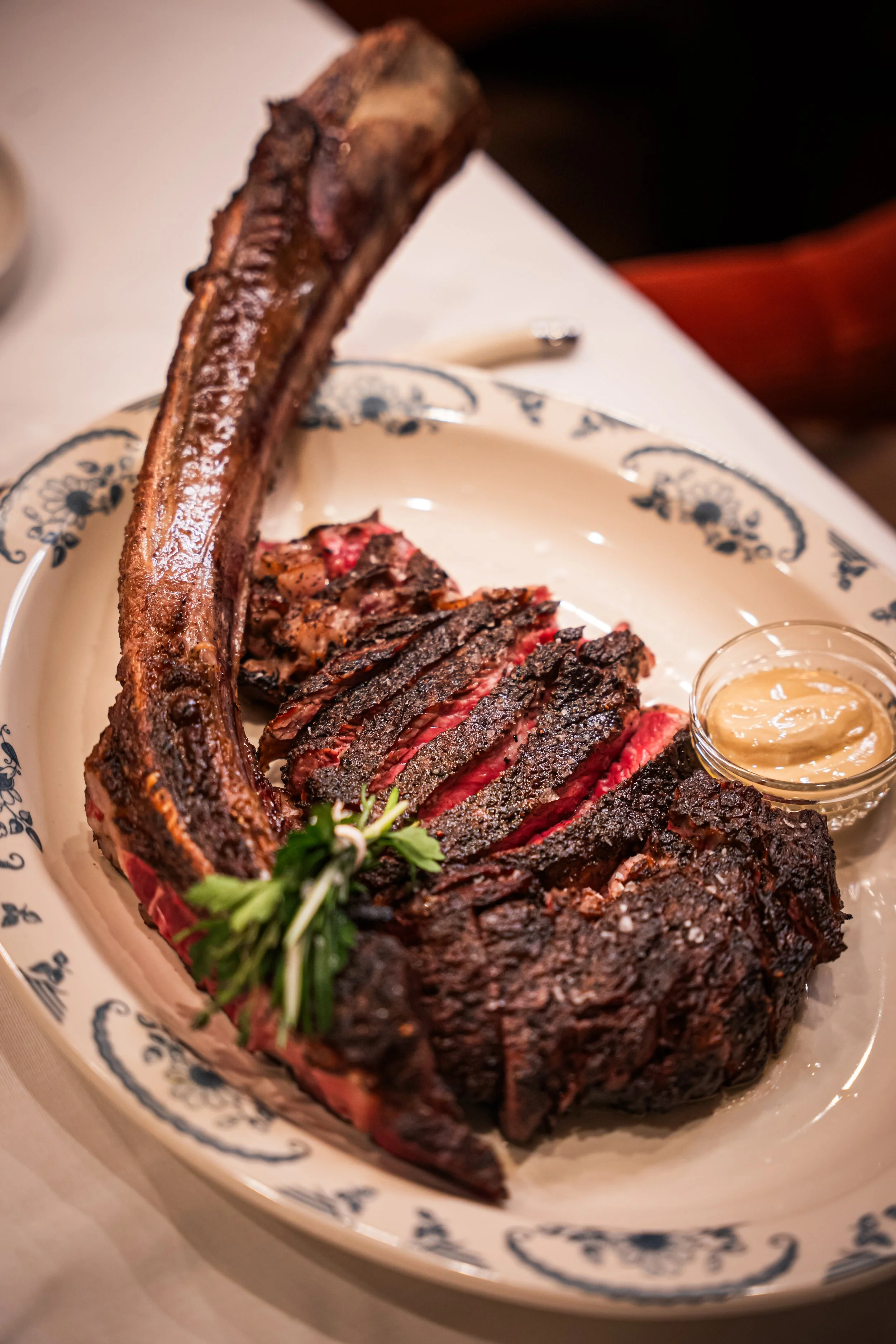 Cooked beef brisket with a long bone, sliced to show a pink center, served on a decorative plate with a small container of mustard sauce, garnished with parsley.