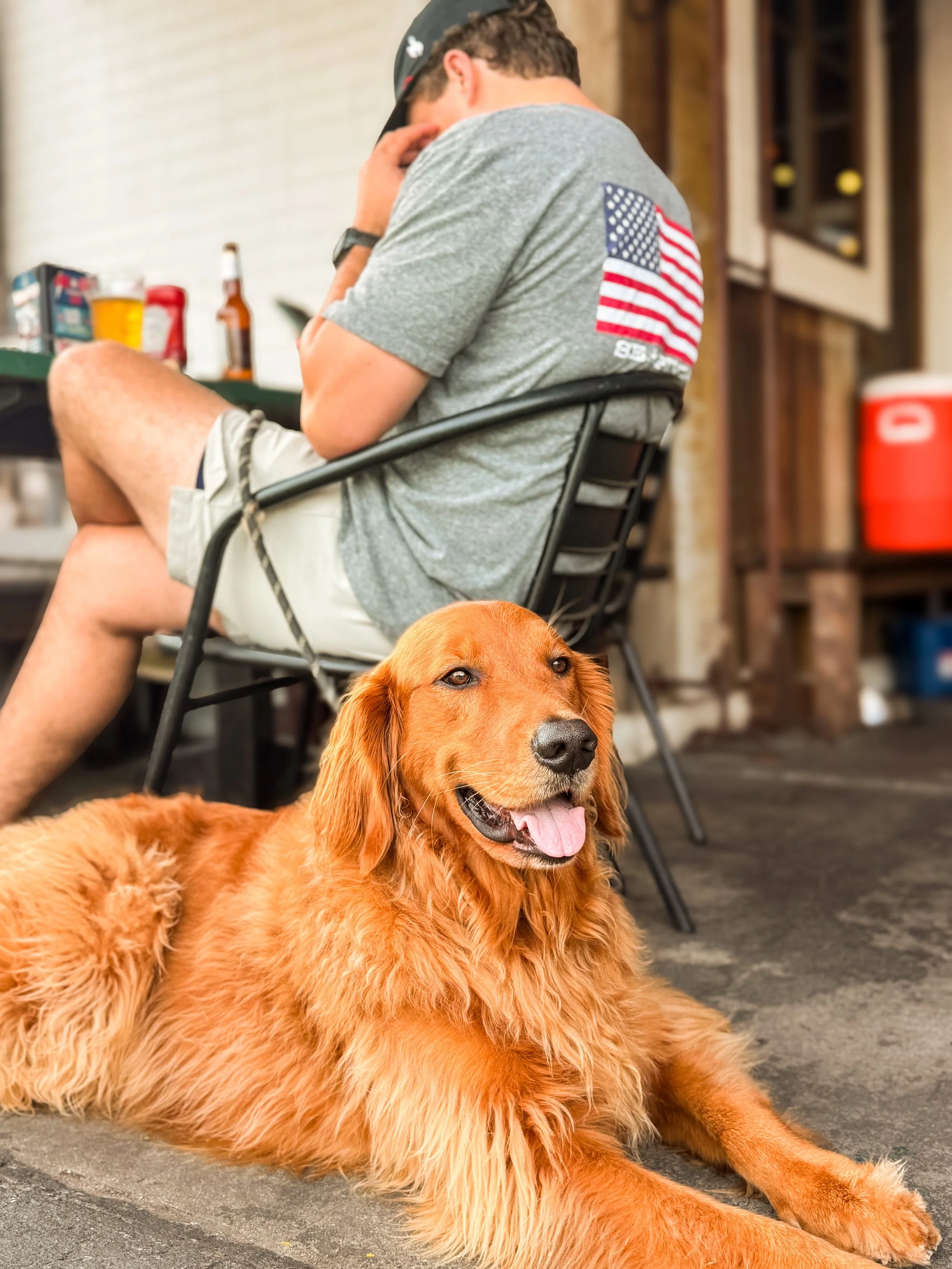 A golden retriever dog lying on the concrete floor with its tongue out, and a man sitting in a chair wearing a gray T-shirt with an American flag patch on the back, in a garage setting.
