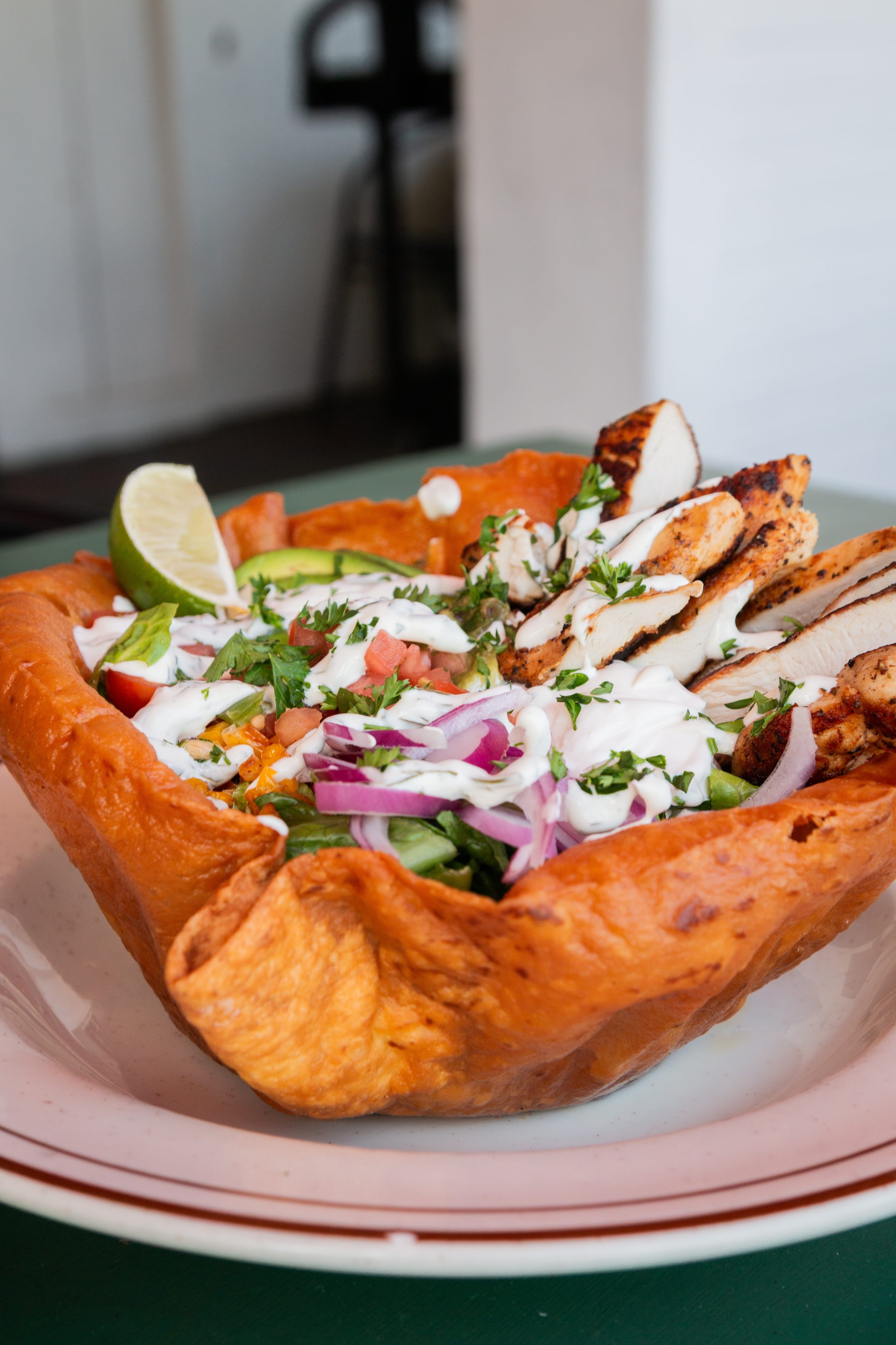 A taco salad served in a crispy shell bowl with grilled chicken, sliced avocado, lime wedge, chopped onions, tomatoes, shredded cheese, chopped cilantro, and drizzled white sauce.