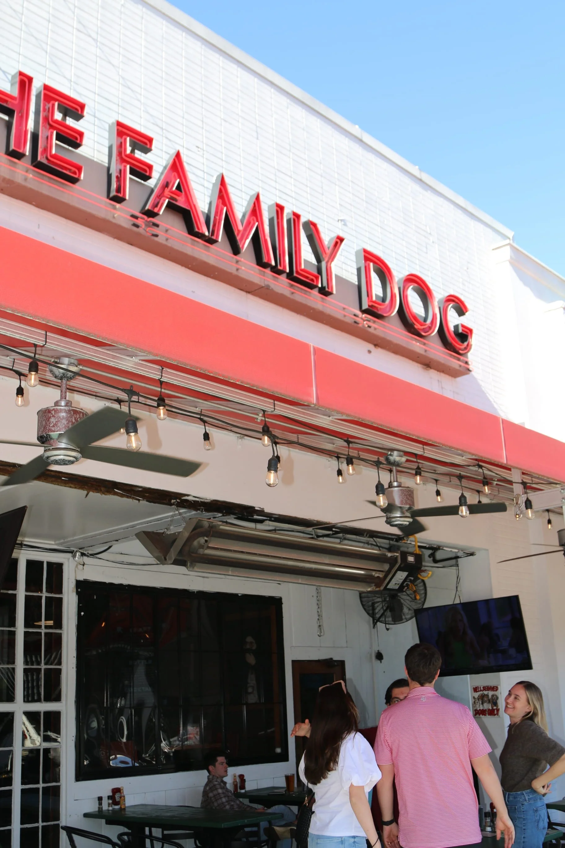 Exterior of a restaurant named The Family Dog with a red awning, outdoor seating, and people standing and talking.