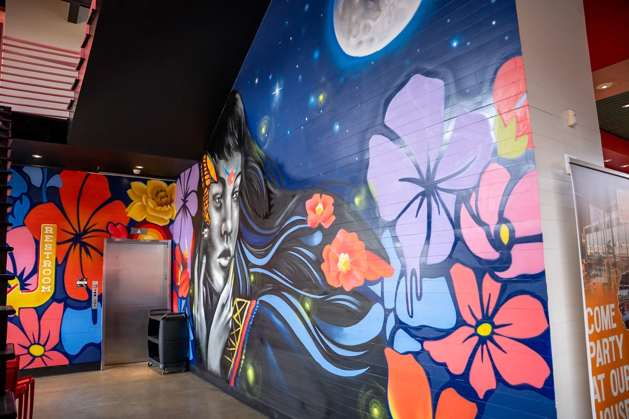 Colorful mural featuring a woman with long black hair, painted in grayscale with vibrant flowers, butterflies, and a moon in the background.