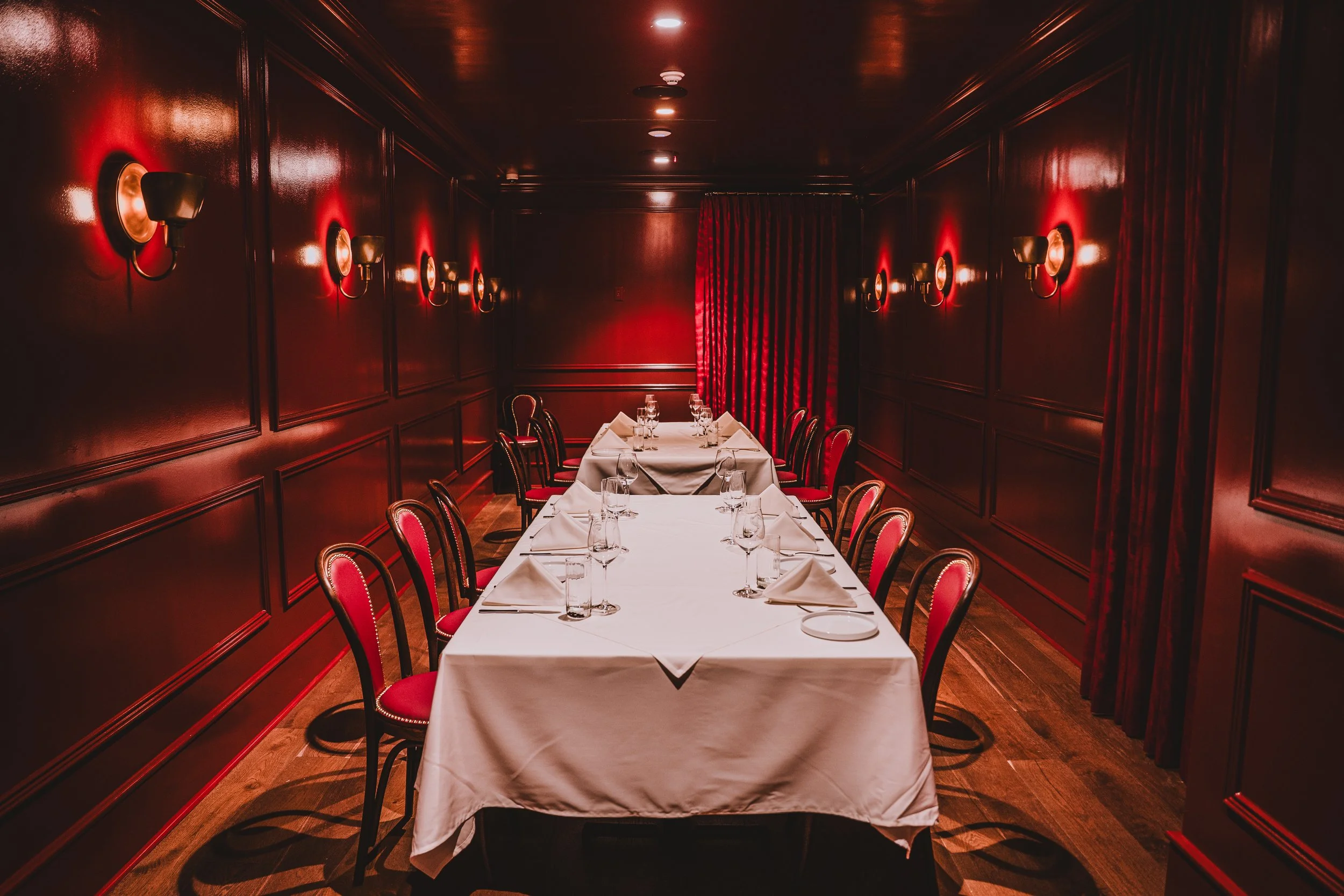 A private dining room with a long rectangular table set with white tablecloths, glasses, and folded napkins. Red upholstered chairs line both sides, and the dark wood-paneled walls have small wall sconces with warm lighting. Red curtains cover the ba
