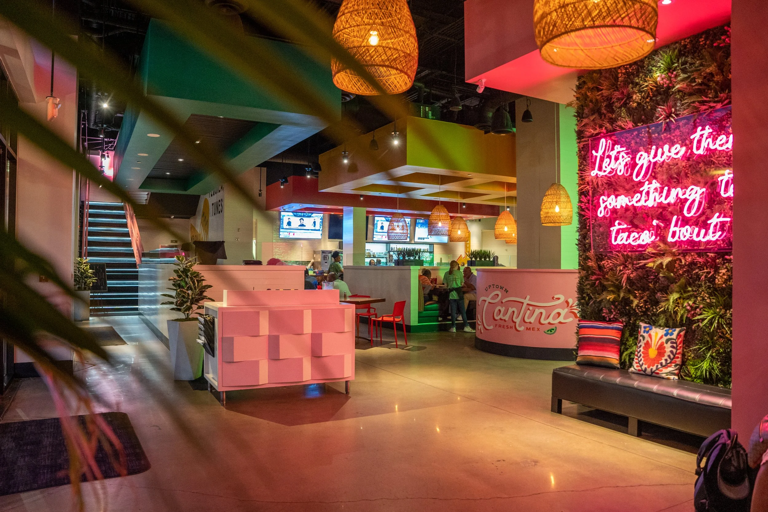 Interior of a colorful restaurant with pink and green accents, hanging rattan lamps, a neon sign with pink text on a floral wall, seating area with a bench and pillows, and multiple TVs displaying news.