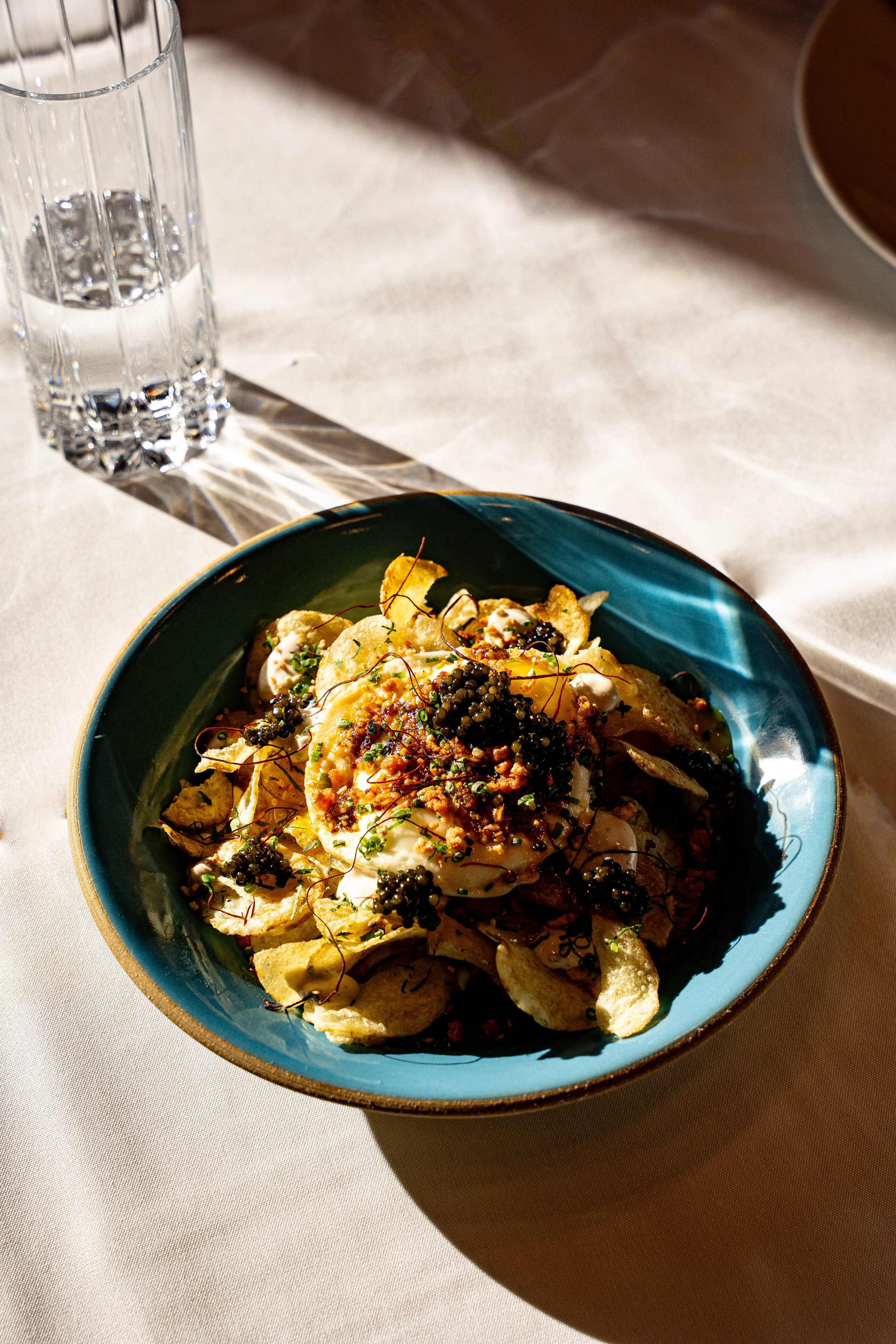 A bowl of chips topped with a poached egg, caviar, and garnishes on a white table with a glass of water nearby.