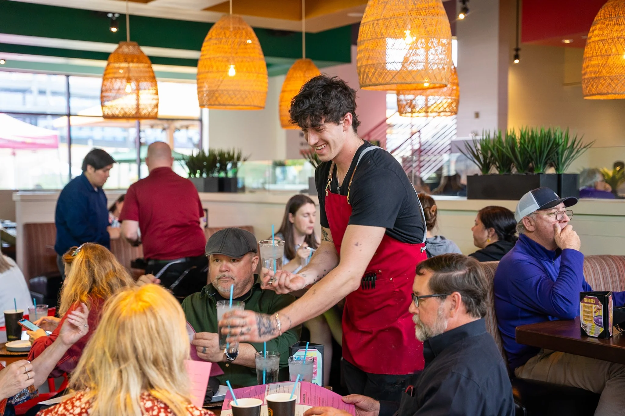 A young man with curly dark hair and tattoos on his arms wearing a black t-shirt and red apron serving drinks to a seated customer in a busy restaurant with warm lighting, green plants, and large woven lampshades.