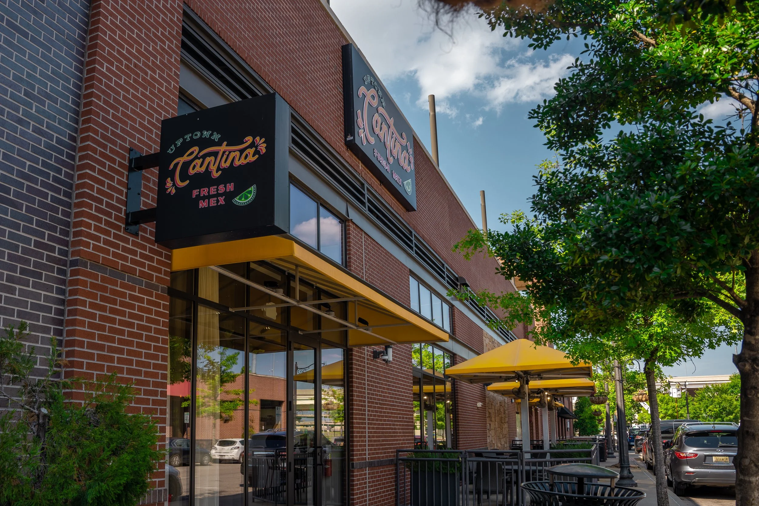Exterior of a restaurant with a black sign that says 'Uptown Cantina Fresh Mex', large glass windows, yellow umbrellas outside on a sidewalk, and trees lining the street.